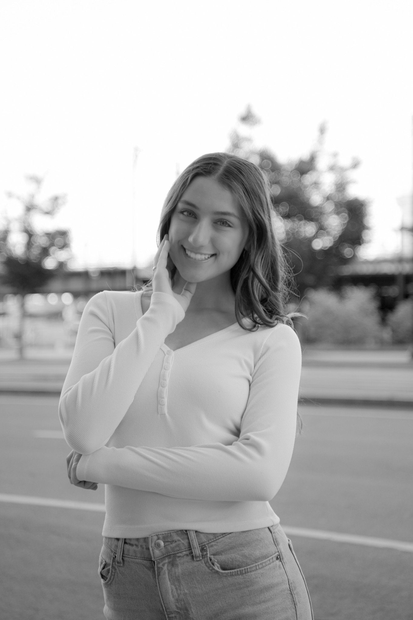 Black and white photo of a young woman smiling outdoors, wearing a long-sleeve top and jeans, with a blurred background of trees and a fence.