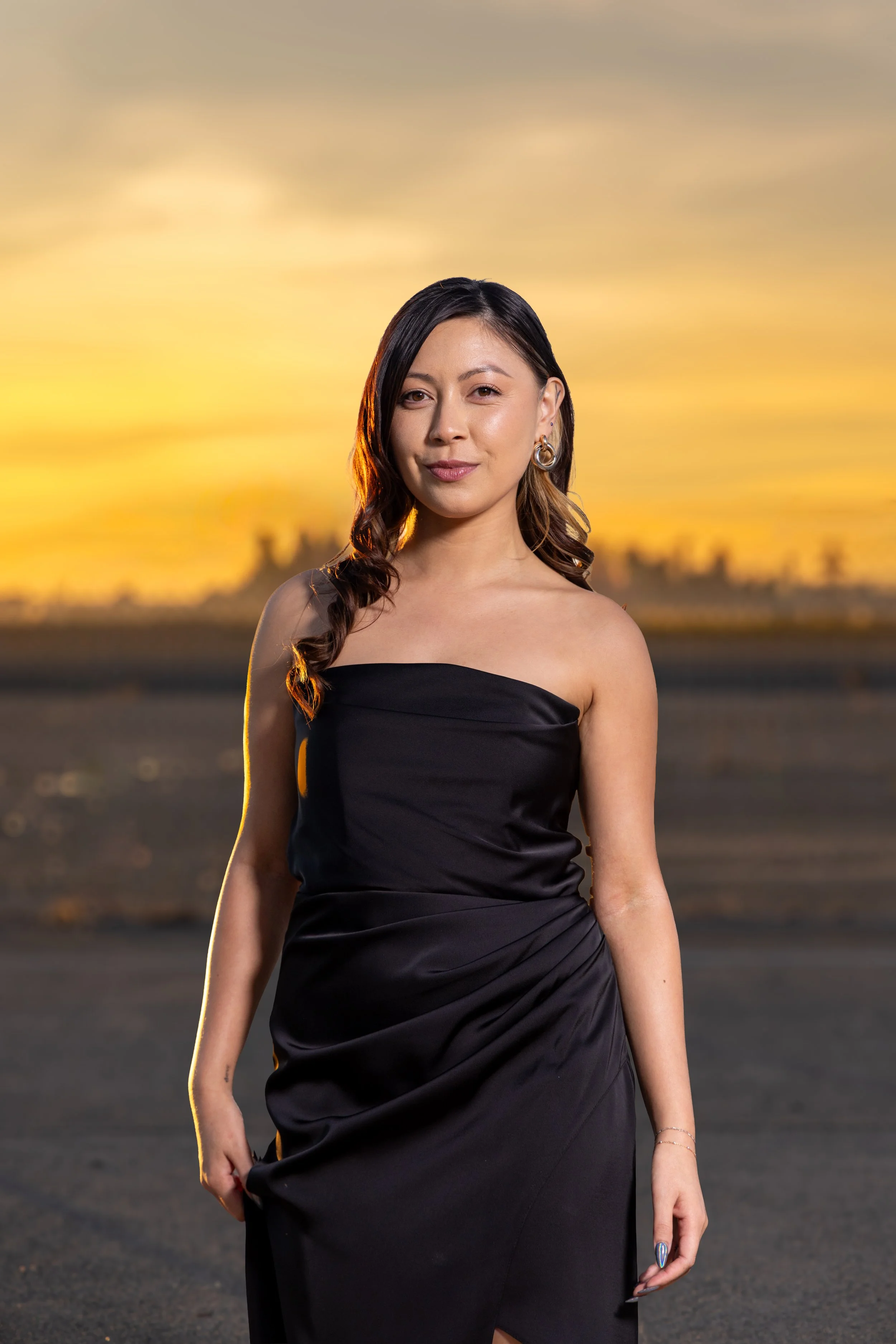 A woman in a black strapless dress standing outdoors at sunset, with a blurred background of a sunset sky and landscape.