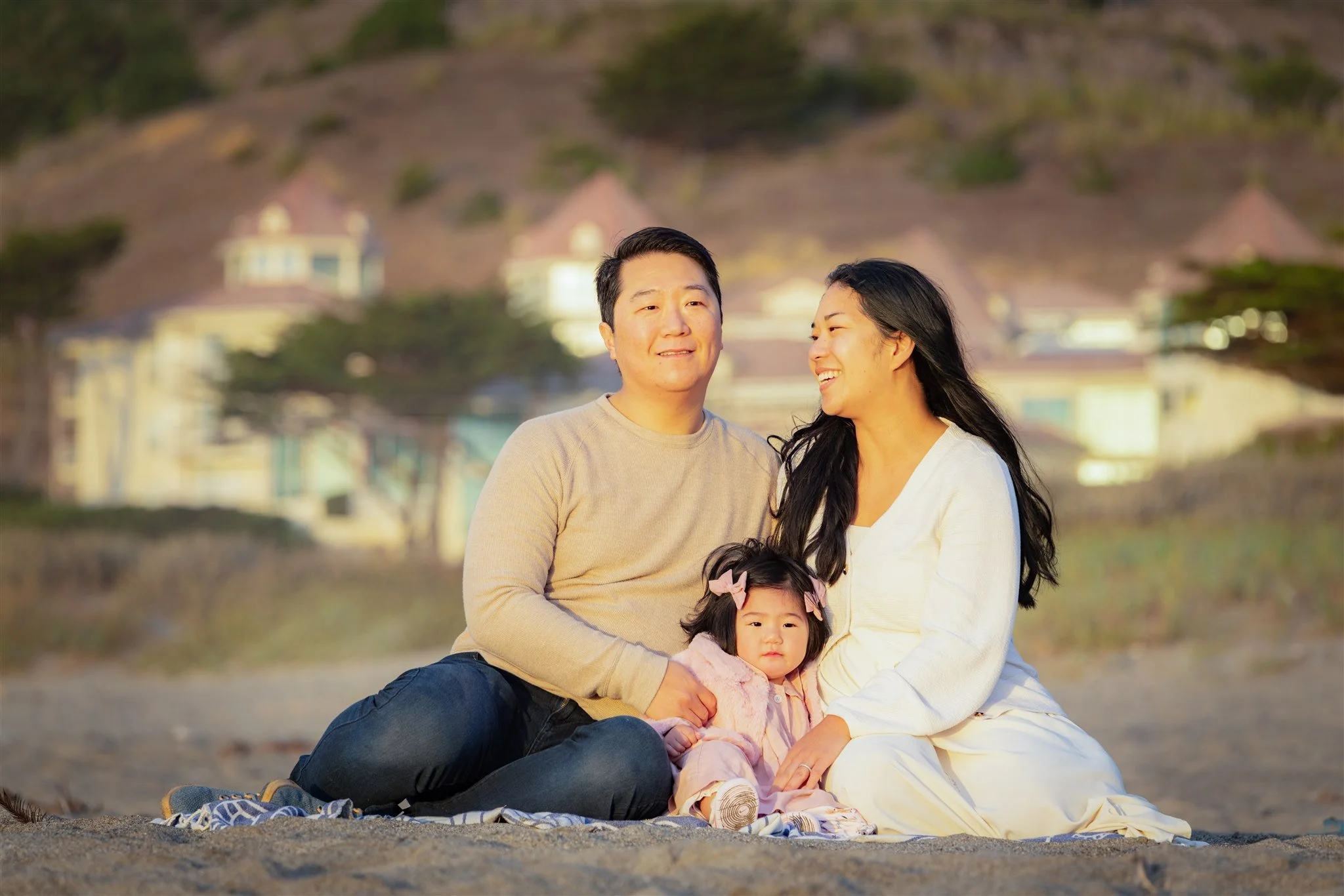 A family of three sitting on a beach with houses and hills in the background, adults smiling and a young girl with pink bows in her hair sitting between them.