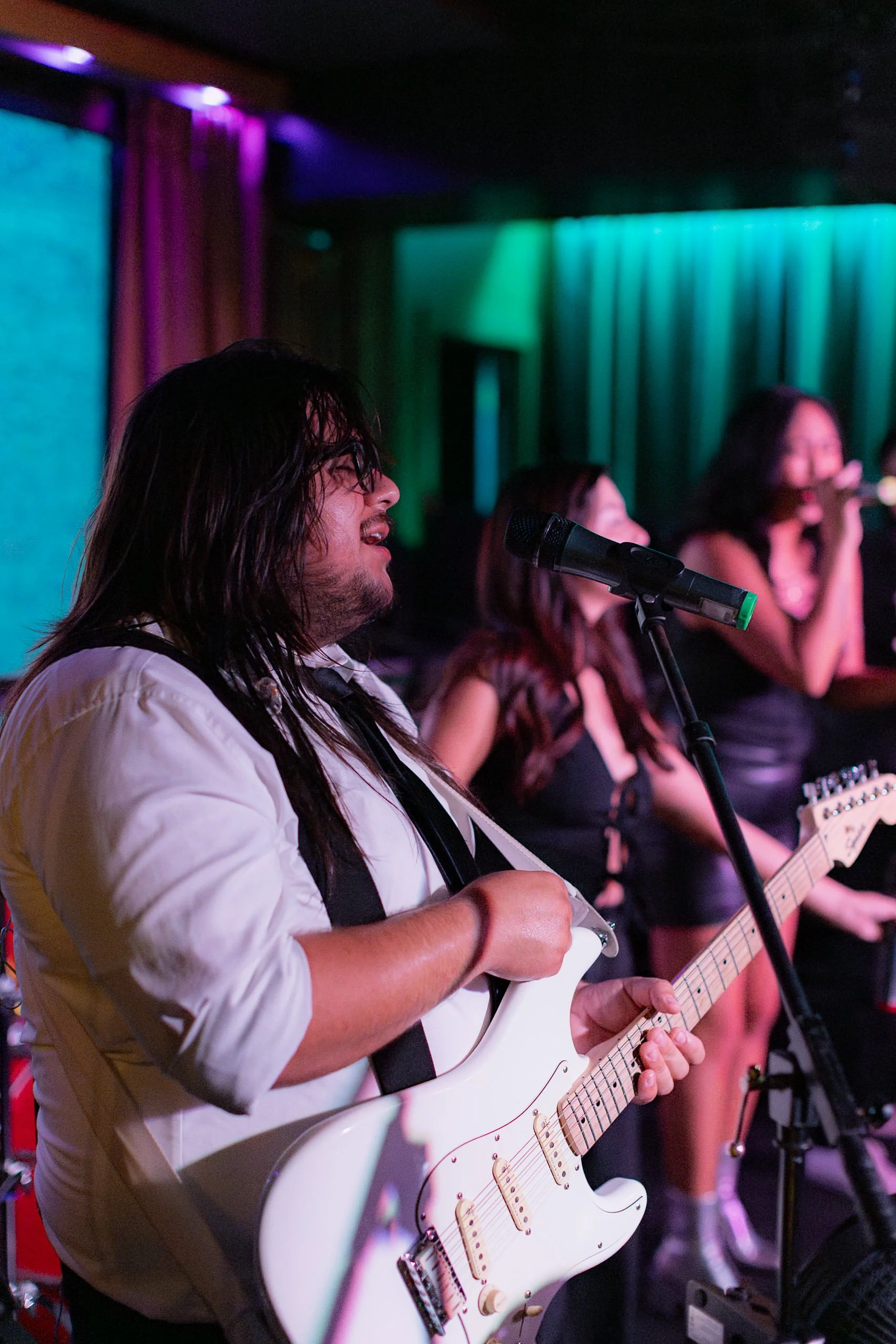 A musician with long hair and glasses playing an electric guitar on stage, accompanied by female singers with microphones, with colorful stage lighting in the background.