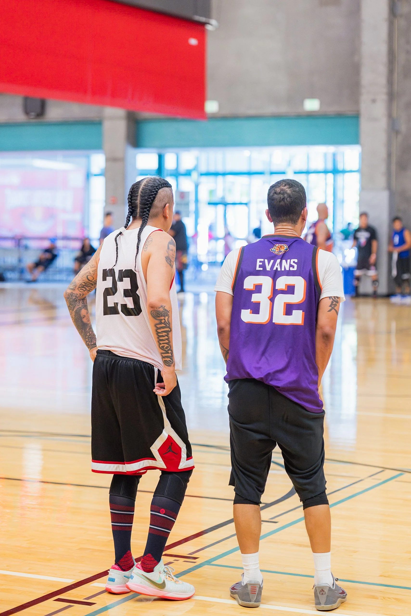 Two basketball players standing on a court, facing away from the camera, one with a white jersey numbered 23, and the other with a purple jersey numbered 32, with the name 'Evans' on the back.