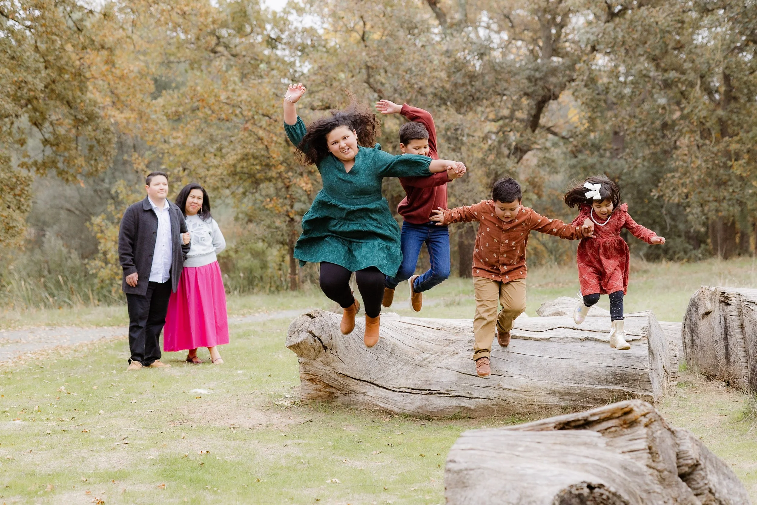 Group of children jumping off a large fallen tree in a park with fall foliage, while two adults stand nearby watching.