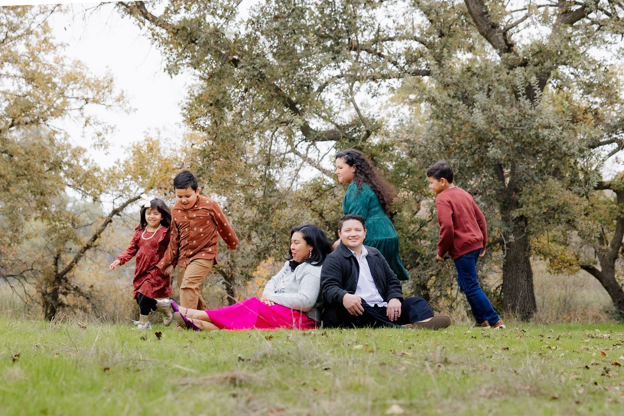 A family of six, including two adults and four children, playing and sitting on the grass in a park with trees and autumn leaves in the background.