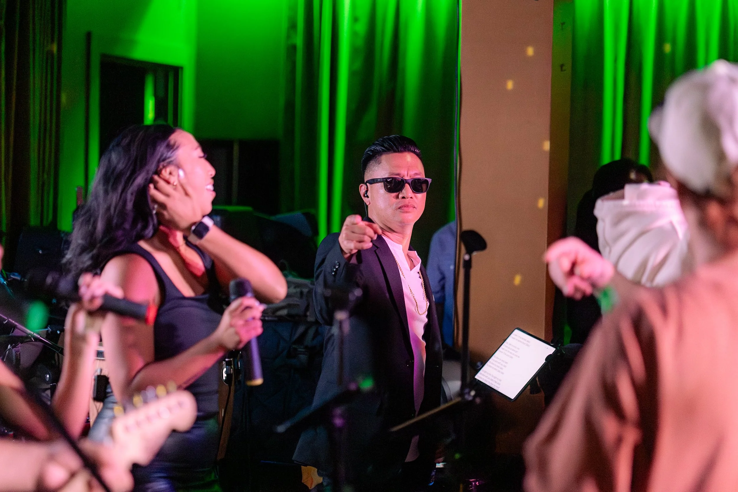 People dancing and singing at a party with green lighting, including a woman holding a microphone and a man wearing sunglasses pointing.