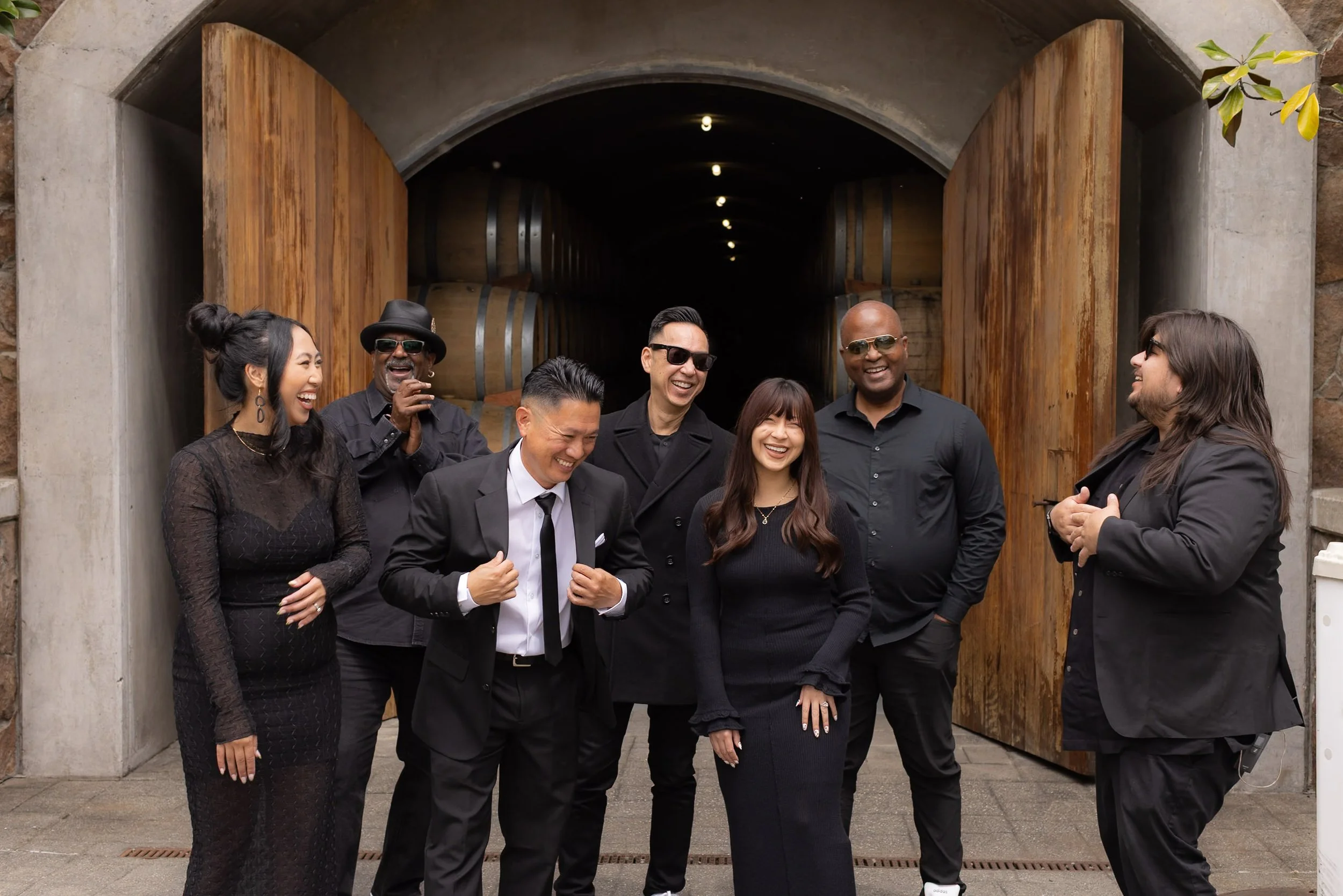 A group of seven diverse people dressed in black, smiling and laughing together in front of a winery entrance with large wooden doors and wine barrels inside.