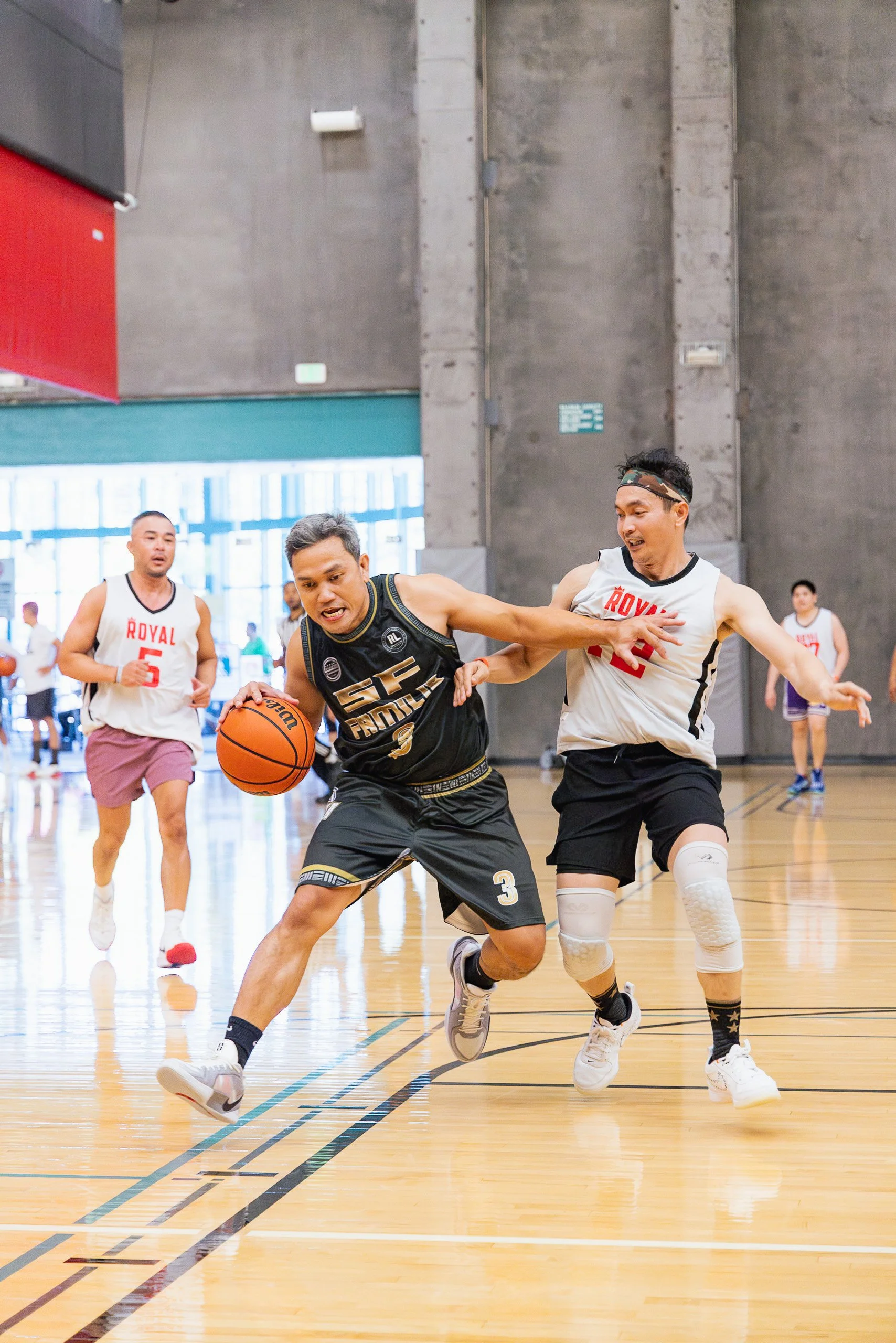 A basketball player in a black jersey dribbling the ball while being defended by a player in a white jersey. Two additional players are visible in the background on an indoor basketball court.