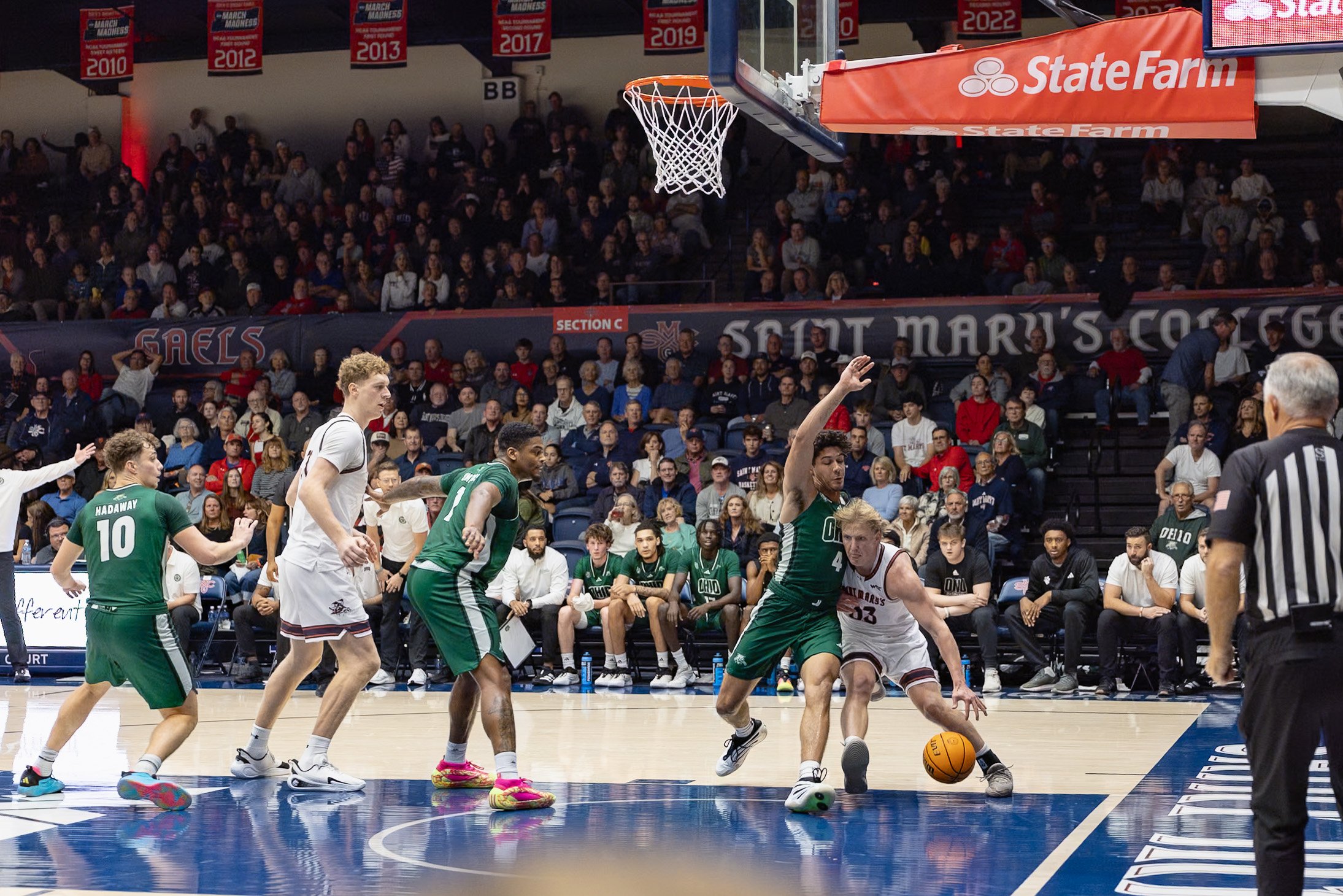 A basketball game in progress with players from two teams competing for the ball near the basket. The players are wearing green and white uniforms, and the game is taking place in an indoor arena filled with spectators.