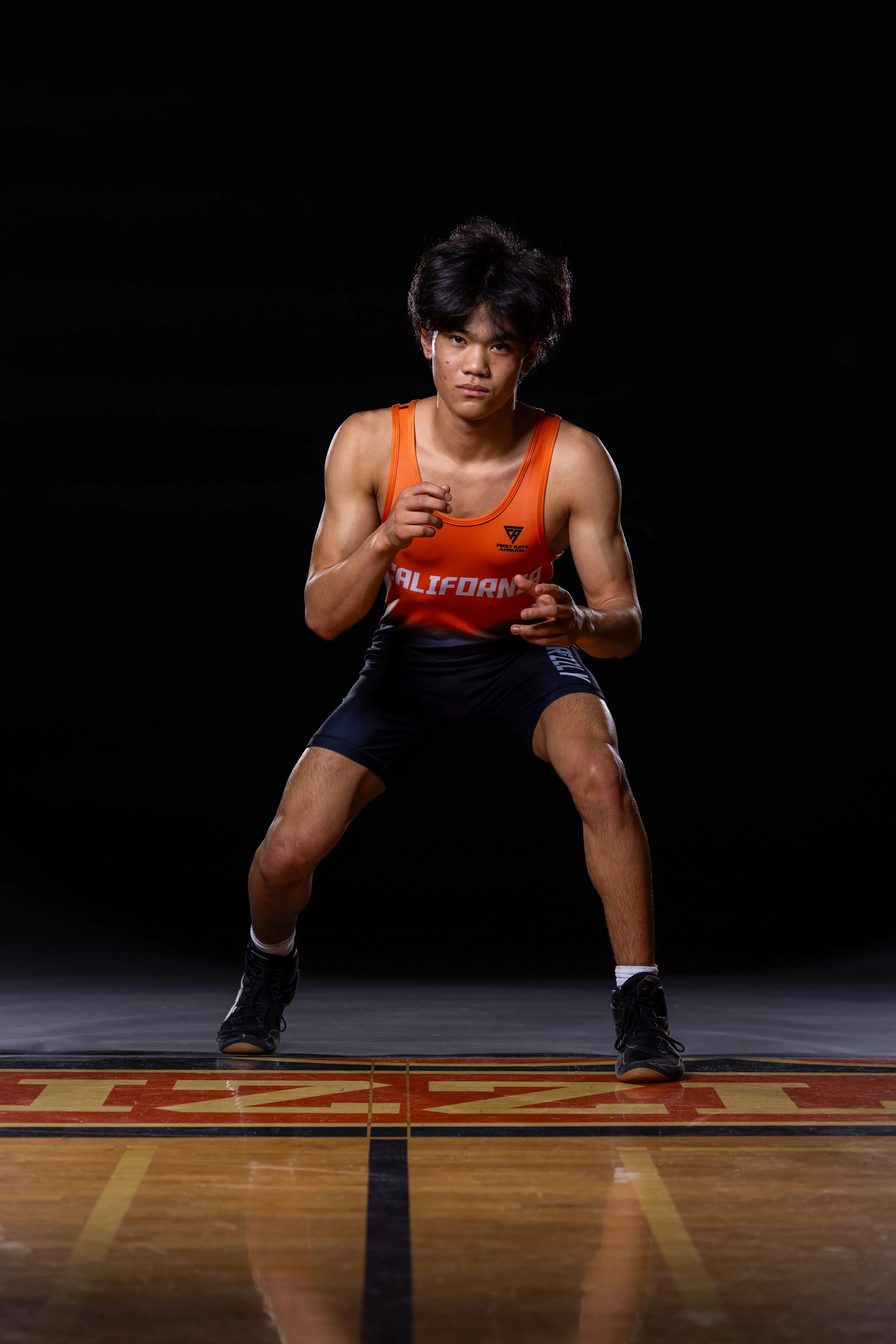 A young male athlete in a ready stance on a basketball court, wearing a red and black uniform with 'California' printed on it, black sneakers, short dark hair, and a focused expression.