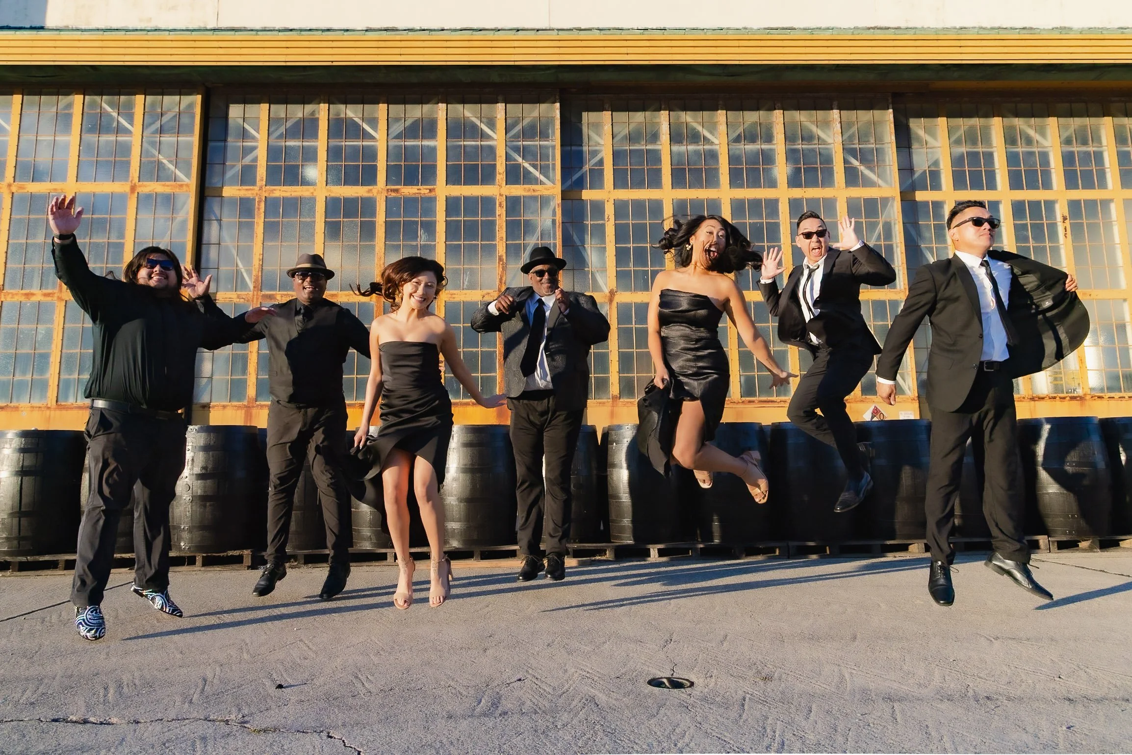 A group of eight people dressed in formal attire jumping in the air outside an industrial building with large windows.