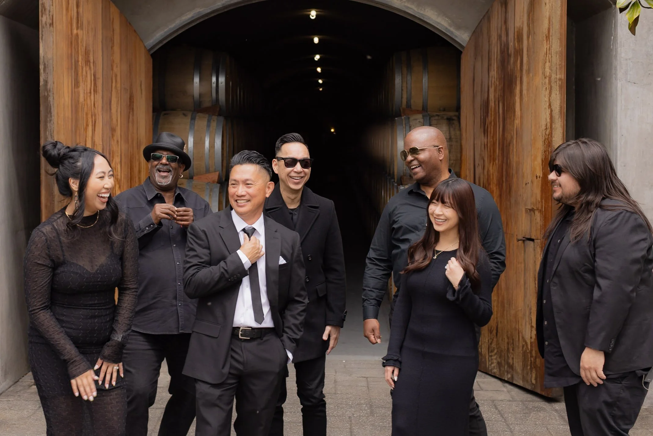 Group of seven diverse people in black attire laughing and smiling together in an urban setting with large wooden barrels in the background.