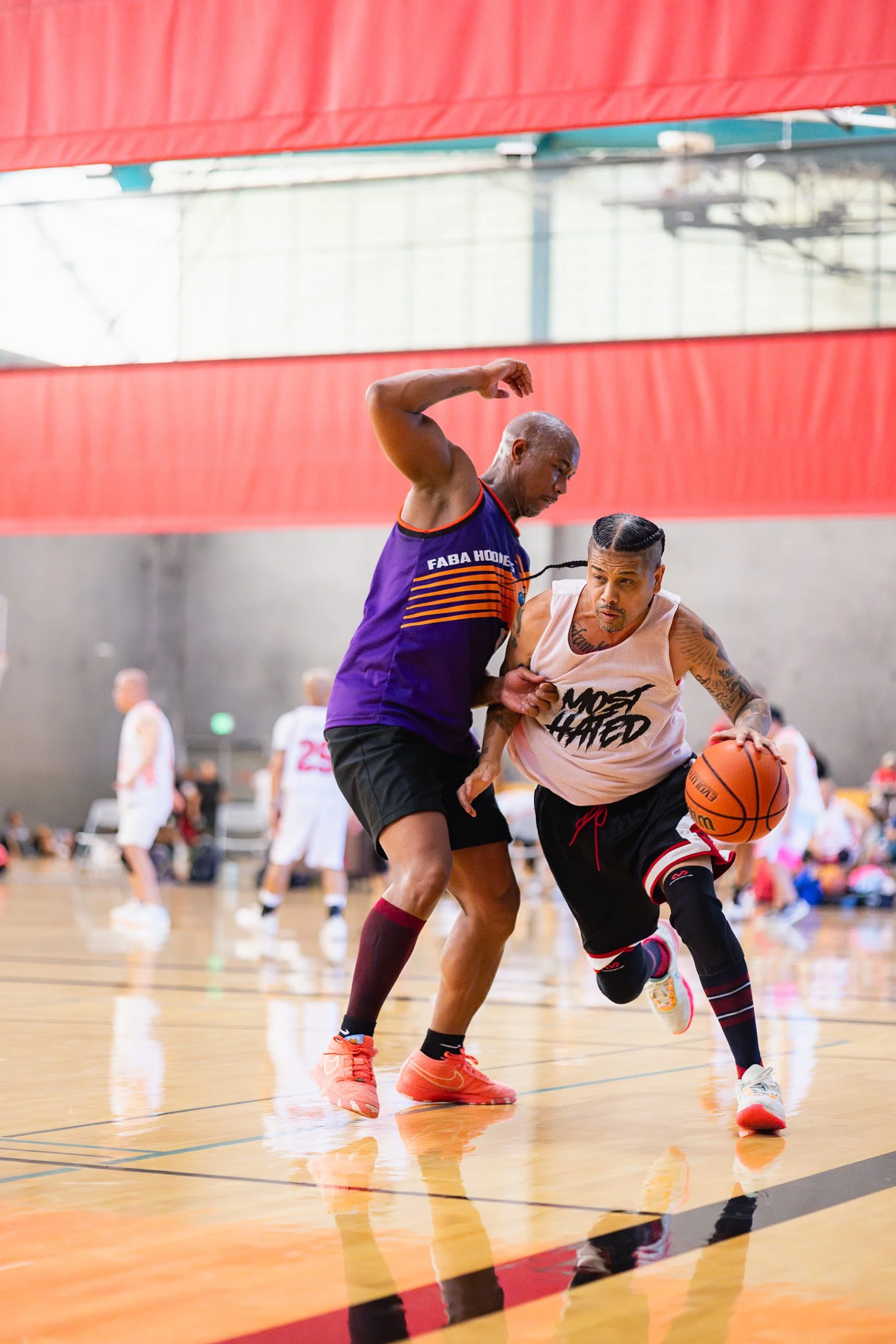Two basketball players in a game: one in a purple jersey defending, the other in a sleeveless white shirt with black and red shorts dribbling the ball on an indoor court.