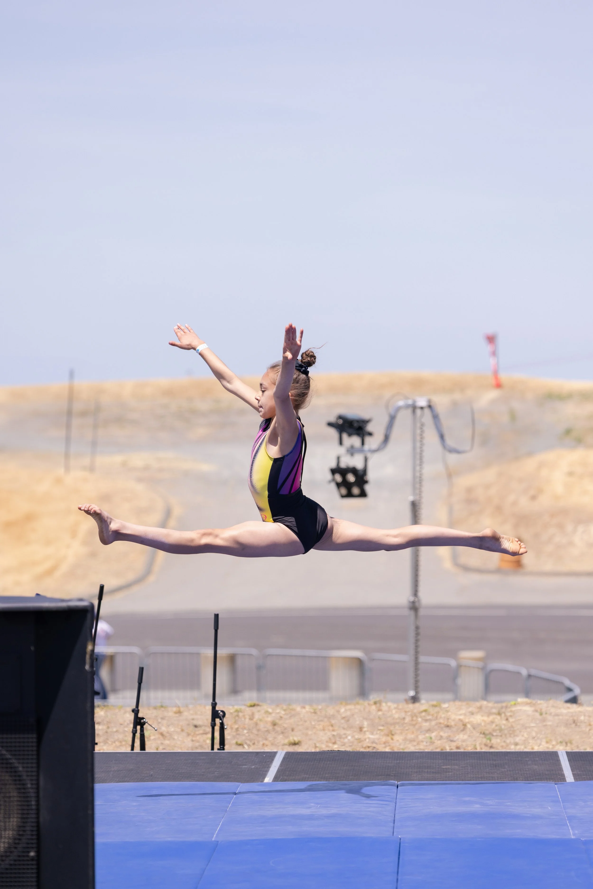 A young female gymnast in a colorful leotard is performing a split leap on a mat outdoors, with a clear sky and dry, hilly landscape in the background.