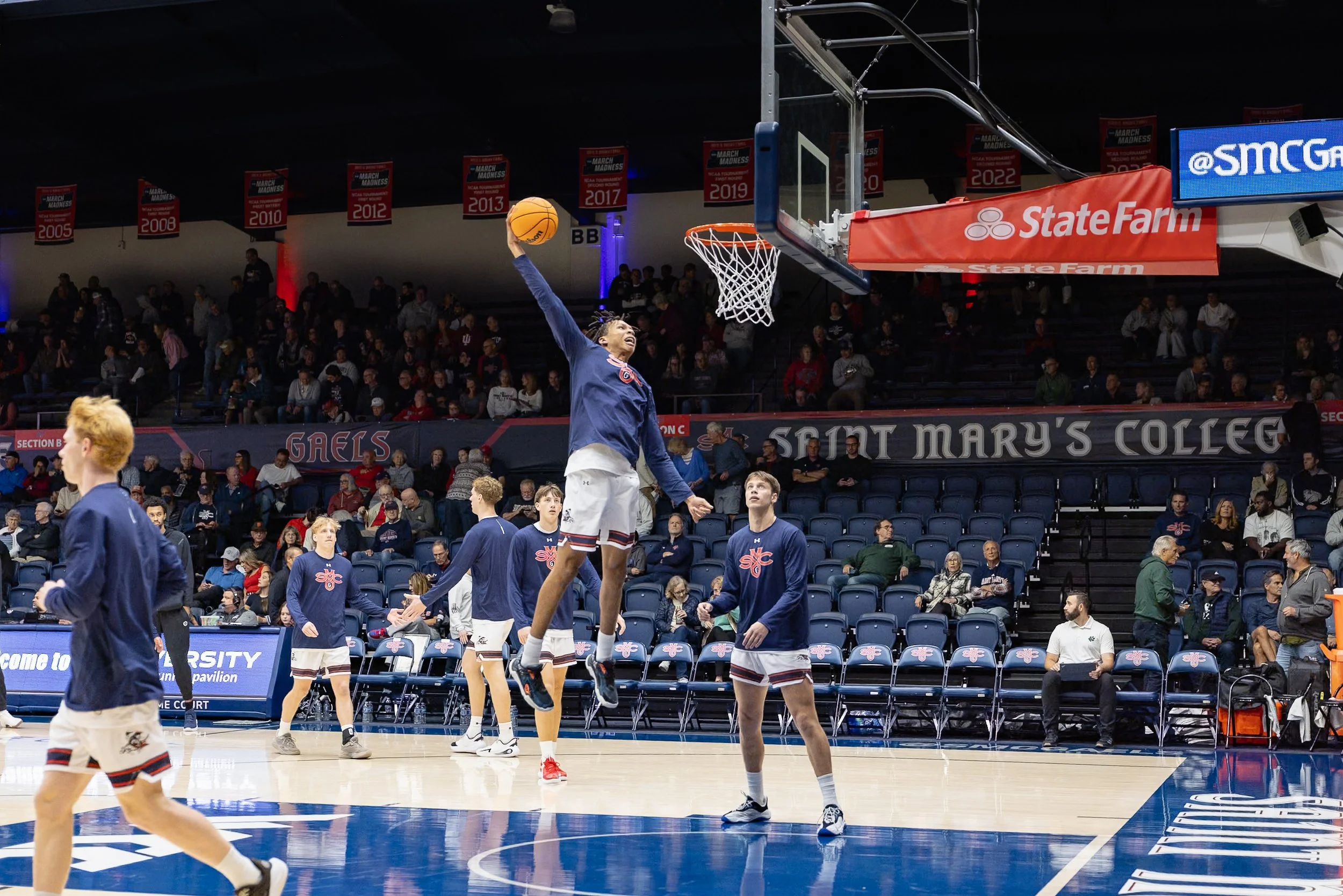 Basketball players warming up before a game at St. Mary's College with some players practicing shots and spectators in the stands.