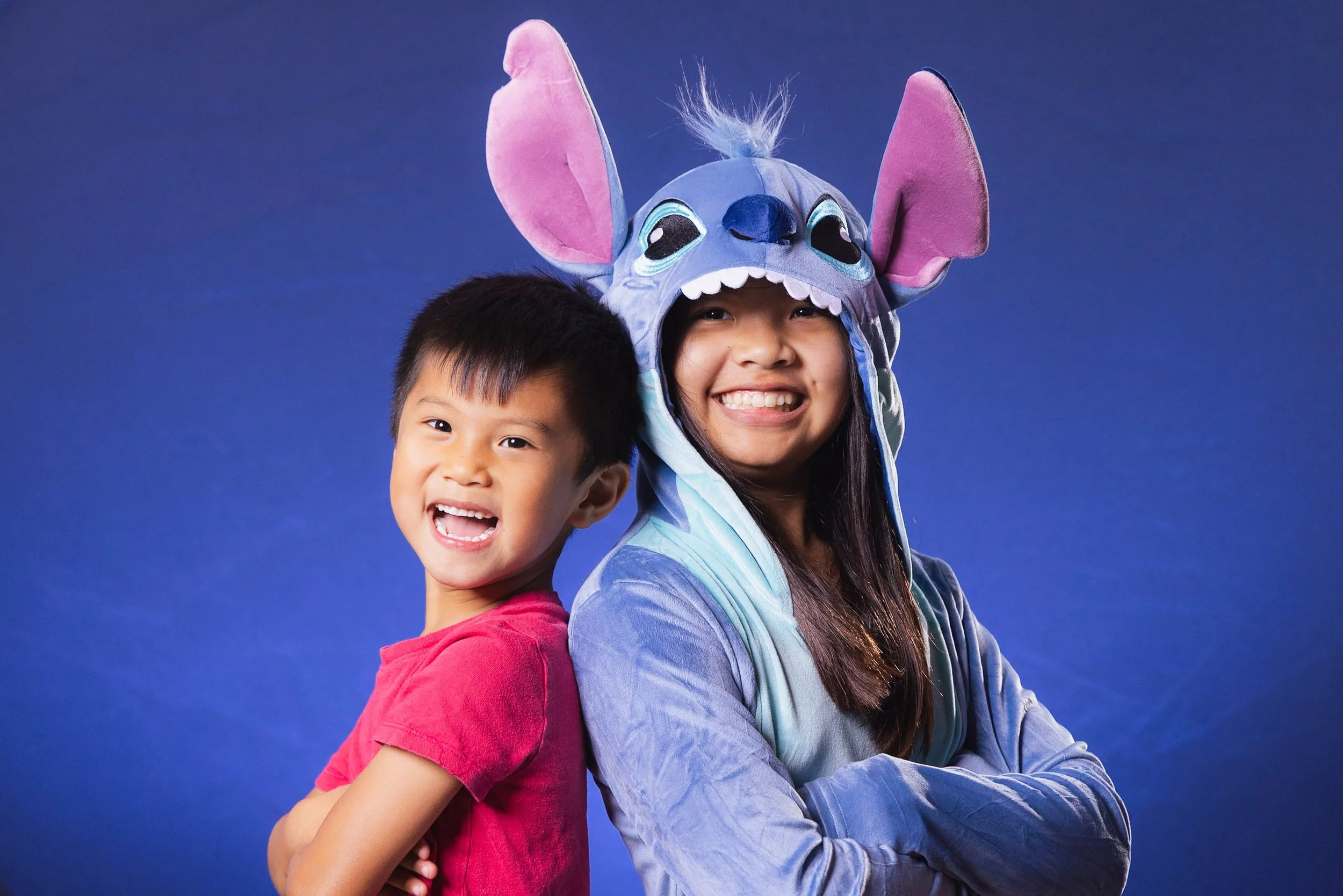 A young boy and girl stand back-to-back smiling. The girl is wearing a Stitch costume hat from Disney's Lilo & Stitch, featuring large ears and a face of Stitch, with her arms crossed. The boy is wearing a red shirt, smiling with arms crossed, agains