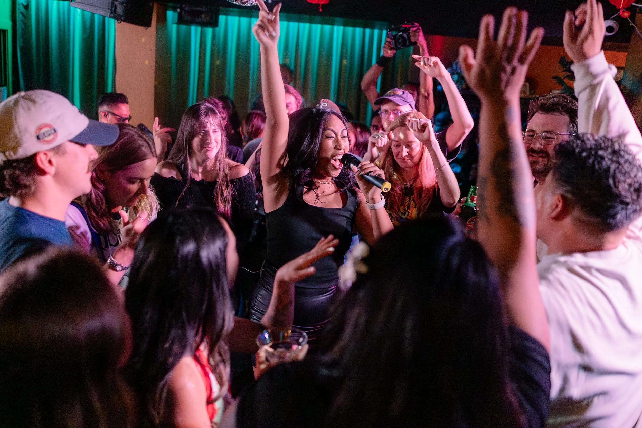 A lively crowd of young adults dancing and singing at a party, with a woman holding a microphone in the center, surrounded by friends in a dimly lit room with green and red lighting.