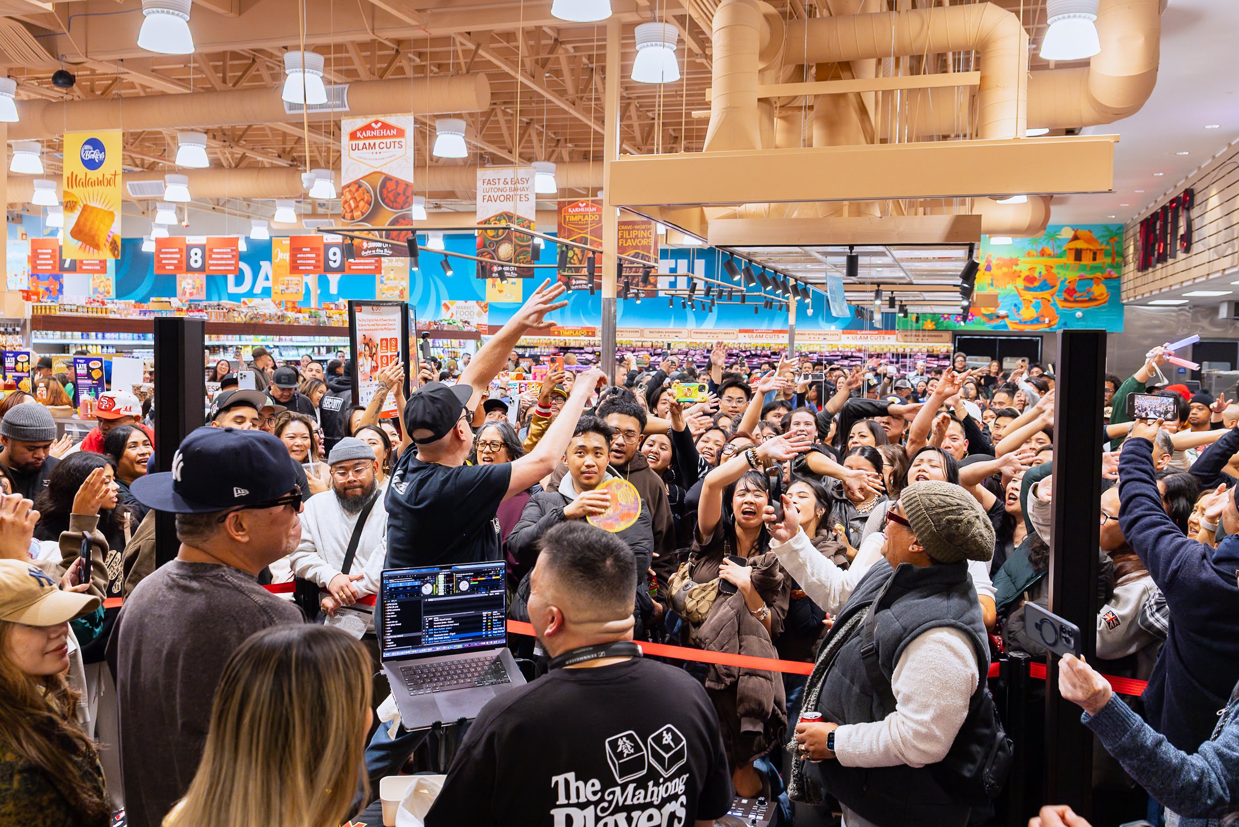 Crowd of people inside a store, gathered around a DJ booth, with some raising their hands, taking photos, and smiling.