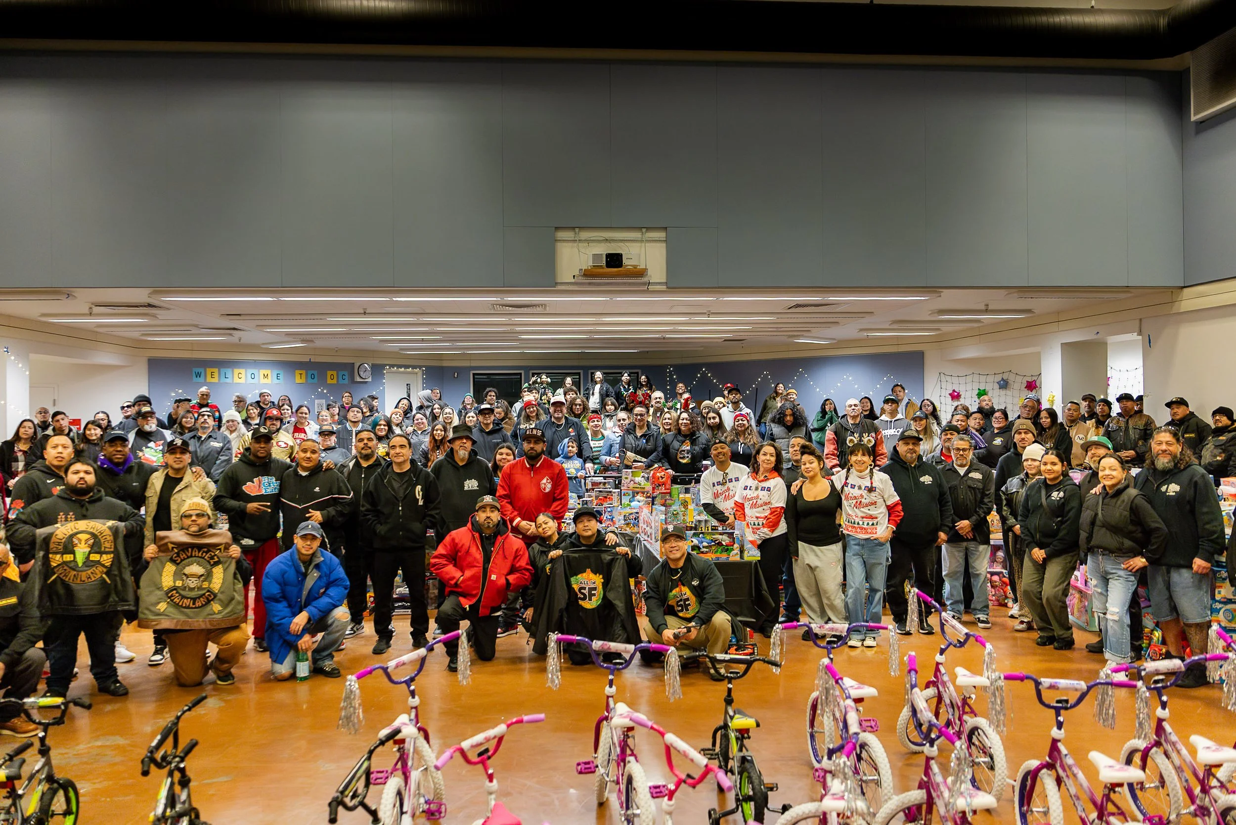 Large group of people gathered indoors for a community event, with festive decorations and numerous toys and bikes in the foreground.