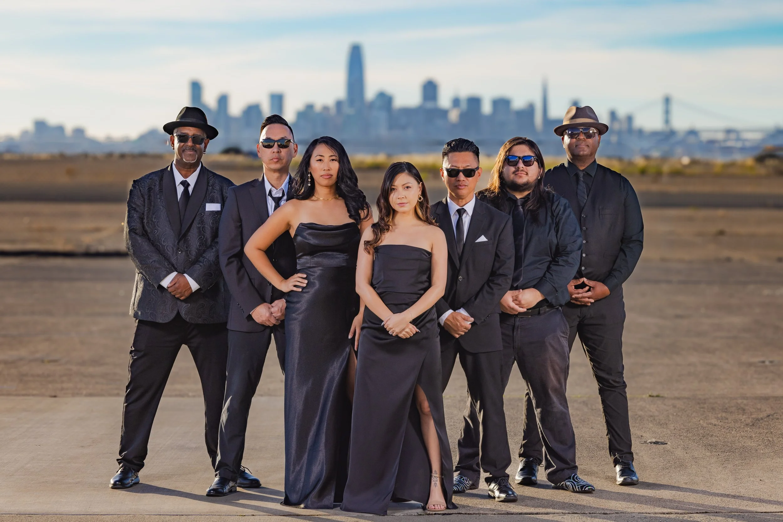 Group of seven people dressed in formal black attire, standing outdoors on a concrete surface with a city skyline in the background, including tall buildings and a bridge, under a partly cloudy sky.