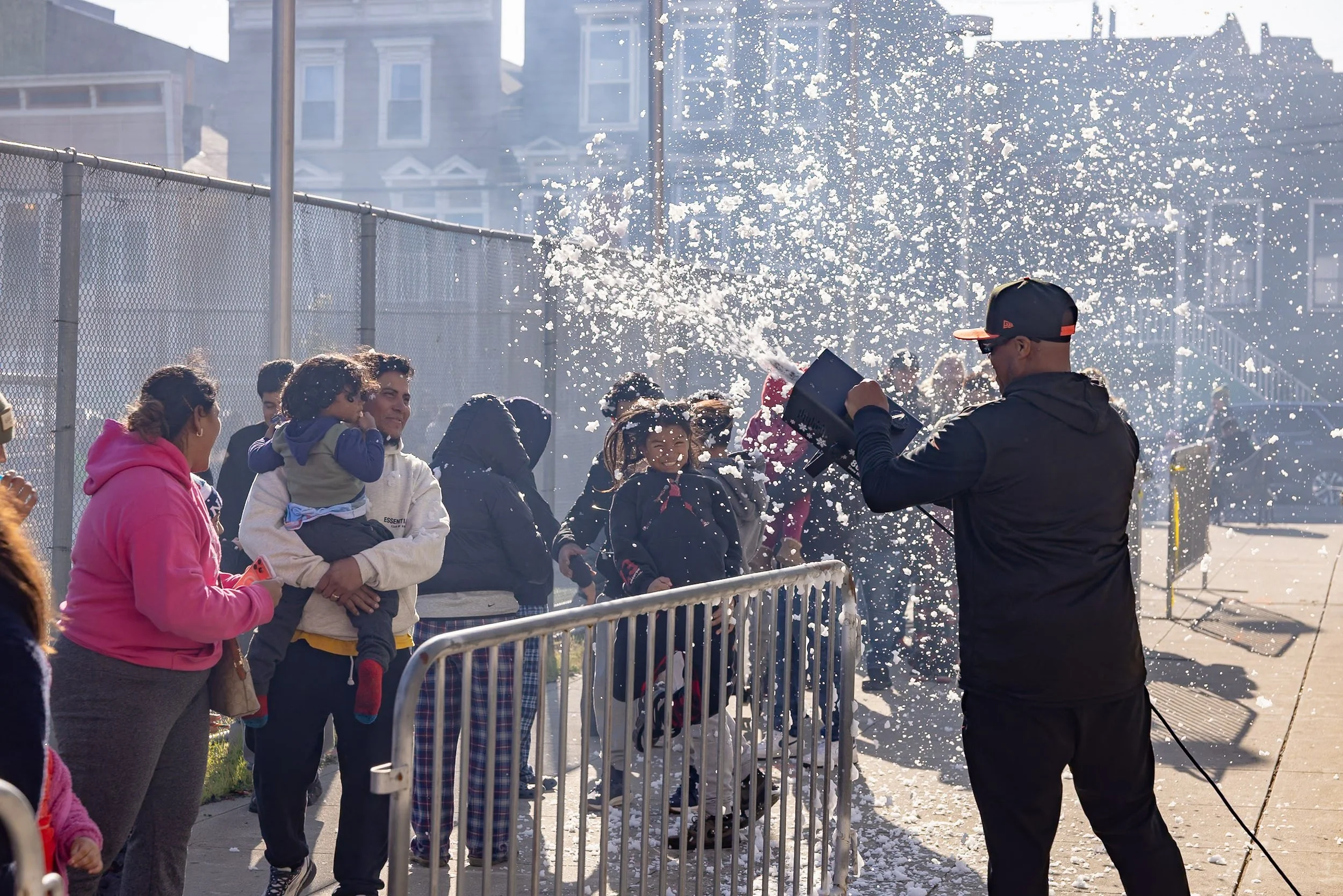 A man sprays foam or bubbles onto a crowd of people, including children and adults, outside on a sunny day to make it seem like it's snowing. Some people are smiling or reacting with happiness and laughter to the foam.
