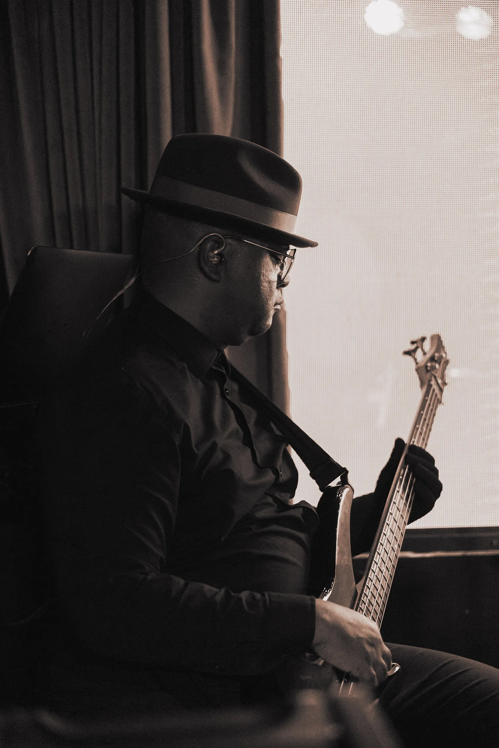 A man wearing a hat and glasses playing an electric bass guitar indoors near a window.