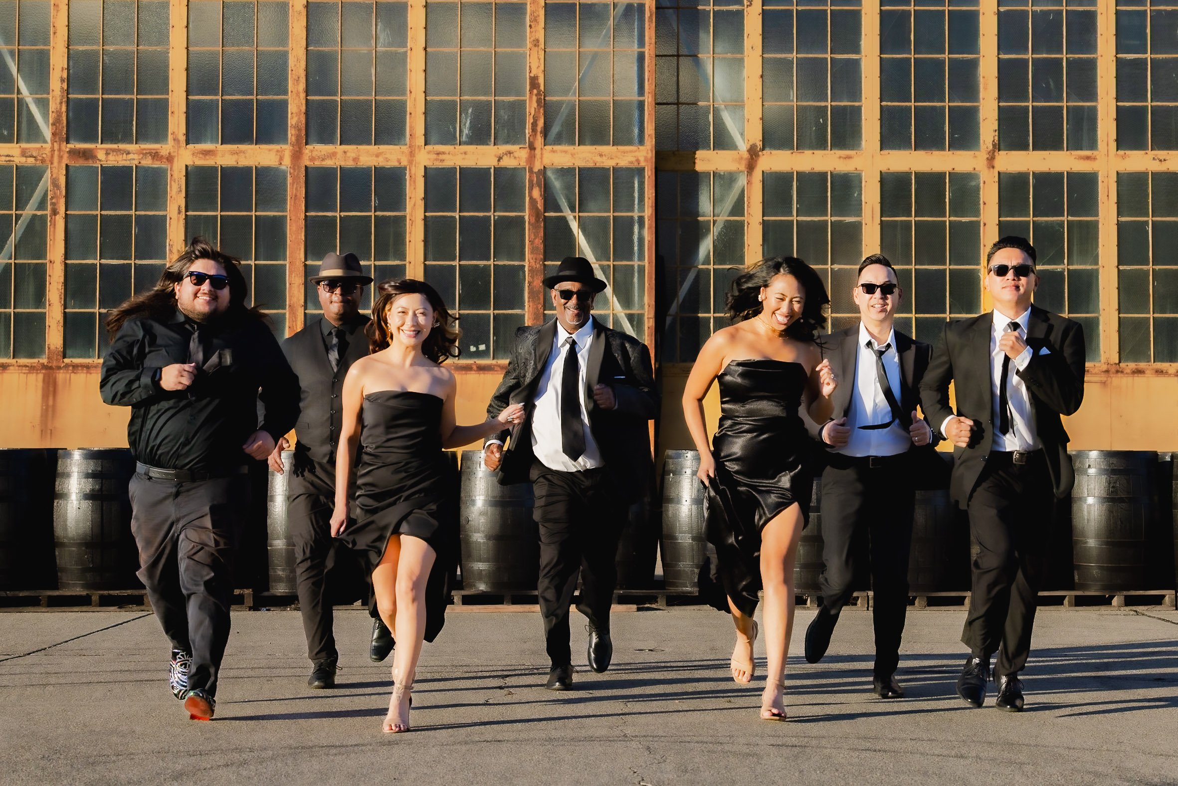 A group of seven people dressed in formal and semi-formal attire running outdoors in front of a rusty industrial building with large windows. The group includes both men and women, some wearing suits and dresses, some wearing sunglasses, all smiling 
