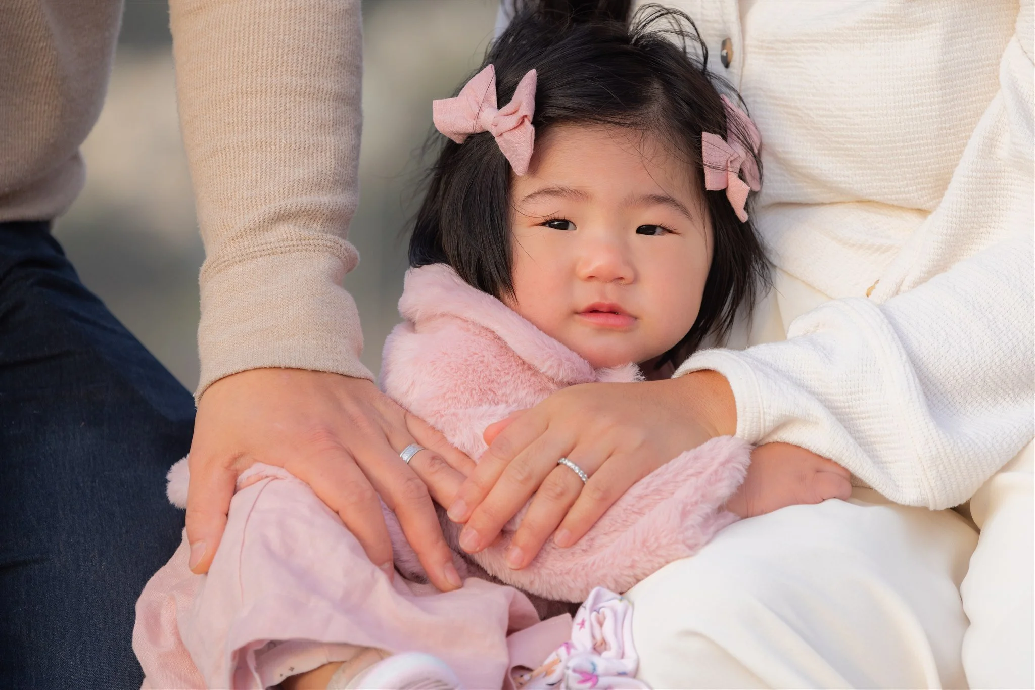A young girl with dark hair and pink bows, wearing a pink fuzzy jacket, sitting on someone's lap while being comforted by two adults, one wearing a light beige sweater and the other wearing a white sweater. The girl looks slightly to the camera with 