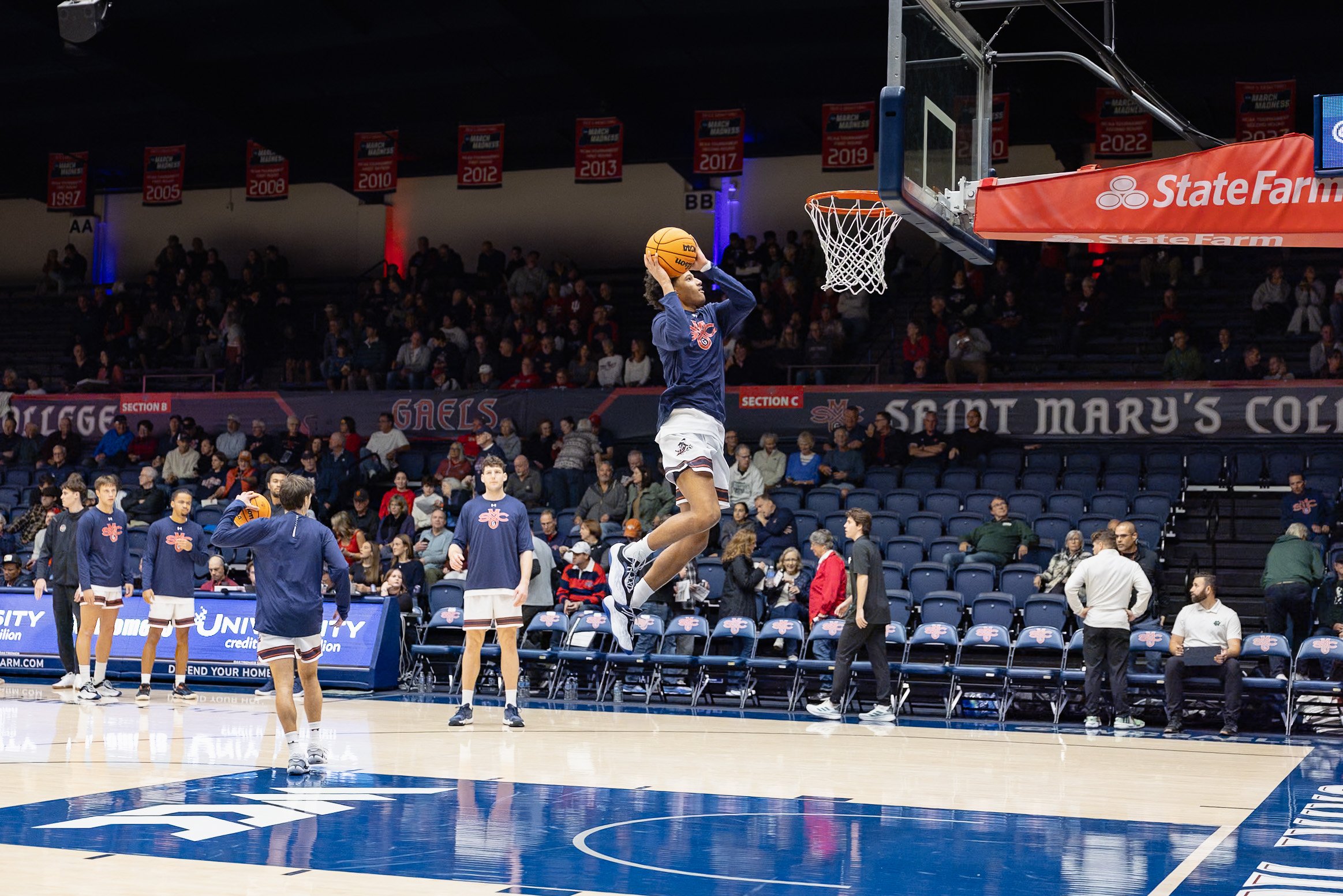 A basketball player in mid-air preparing to shoot during a warm-up on an indoor court, with teammates and spectators in the background.