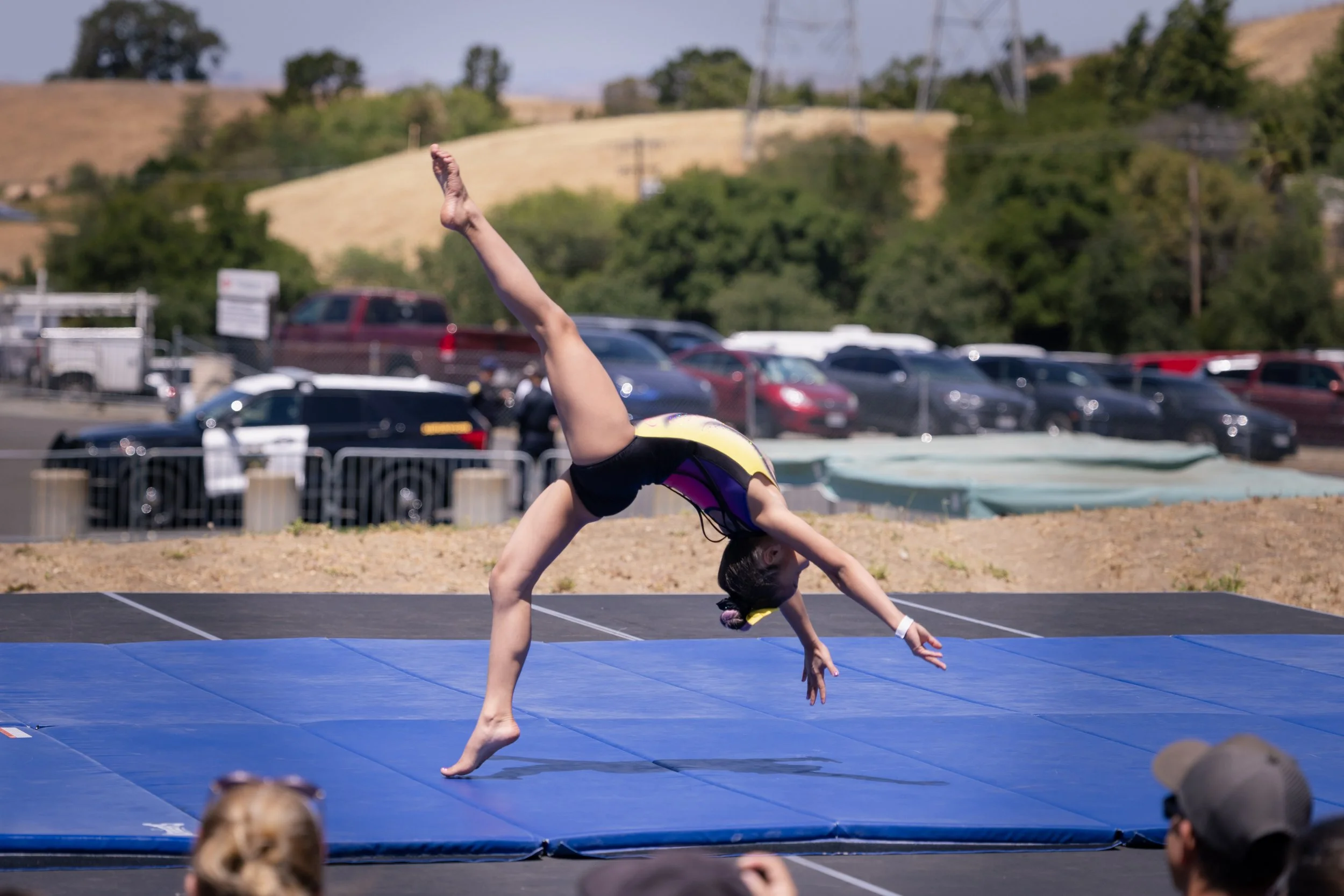 A girl performing a gymnastics move on a blue mat outdoors, with mountains, trees, parked cars, and spectators in the background.