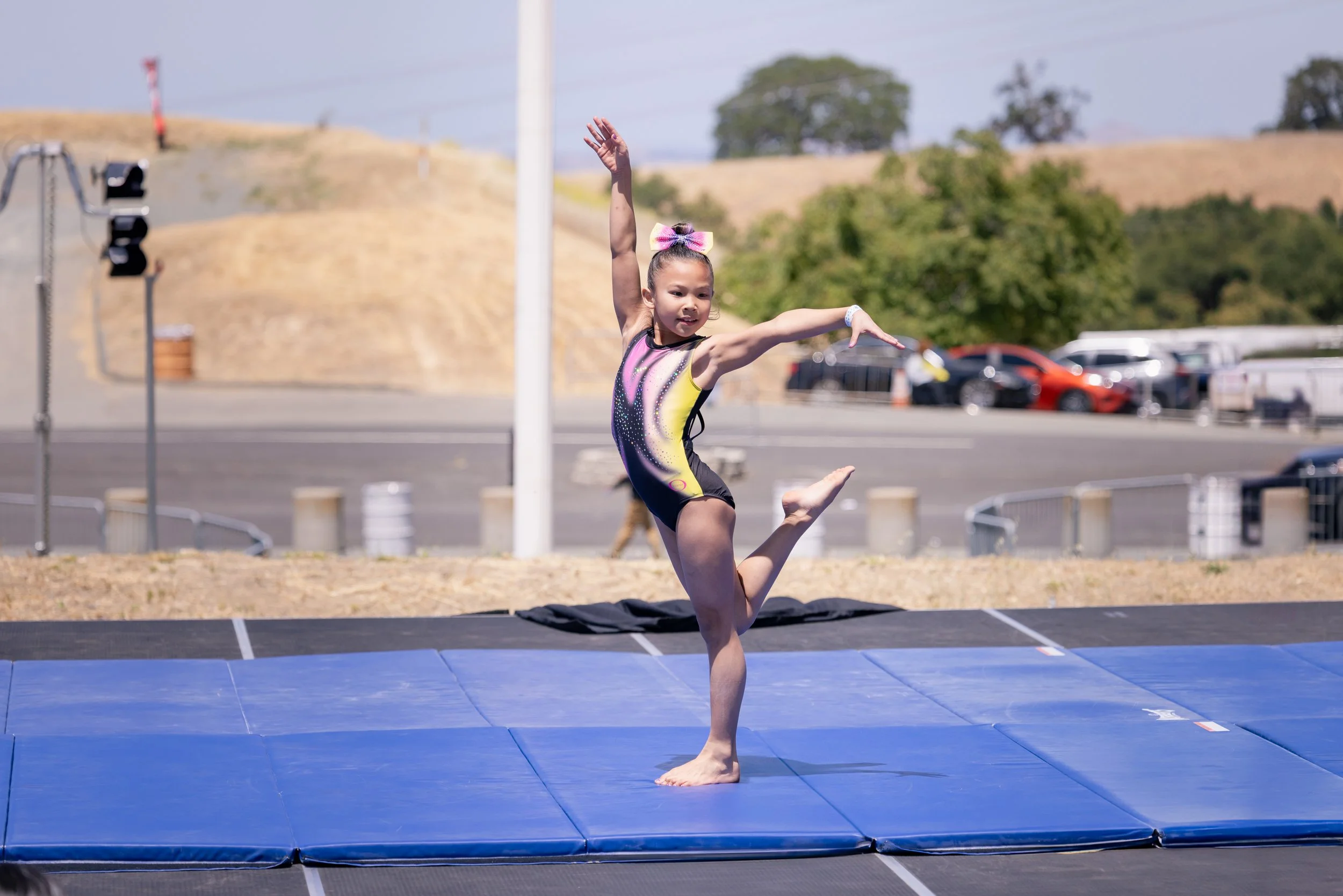 Young girl practicing gymnast routines on a blue mat outdoors, wearing a colorful leotard and a pink hair bow, with her arms raised and one leg lifted.