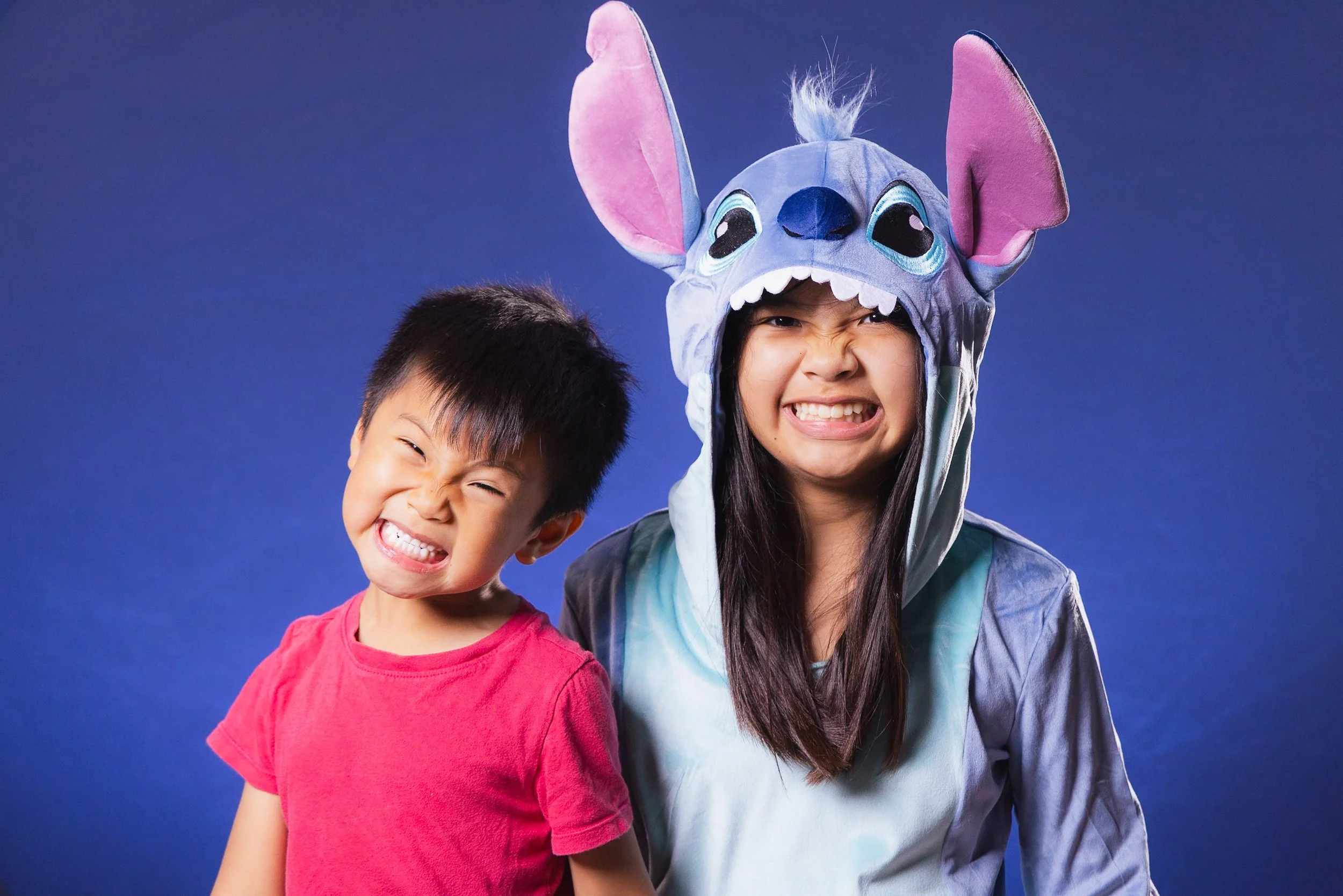 A young boy and girl smiling, the girl wearing a Stitch character costume with large ears and face hood, against a blue background.