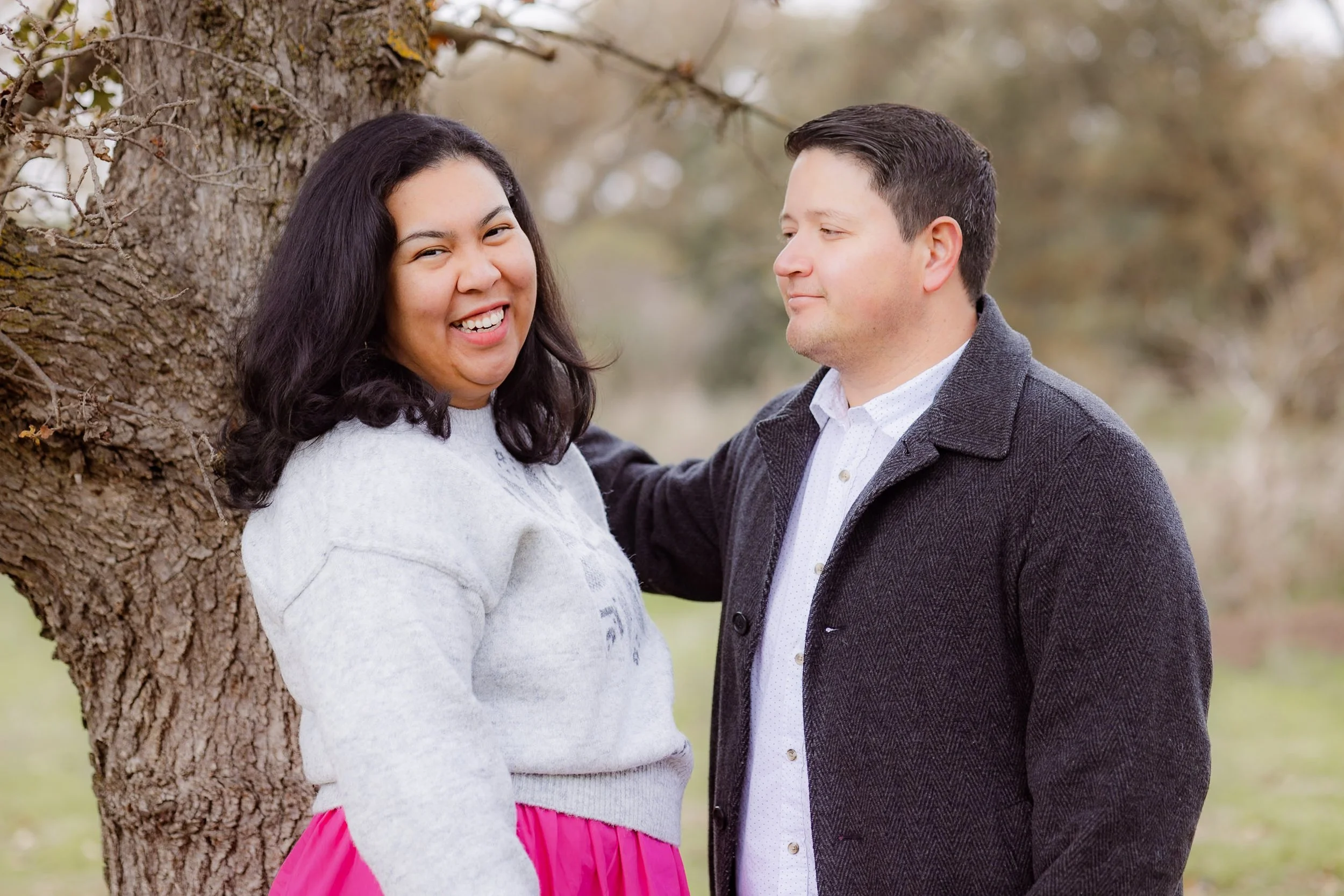 A smiling woman with dark hair wearing a light gray sweater and pink skirt, standing outdoors next to a tree. A man in a dark coat and white shirt stands beside her, looking at her. They are in a natural setting with blurred trees in the background.