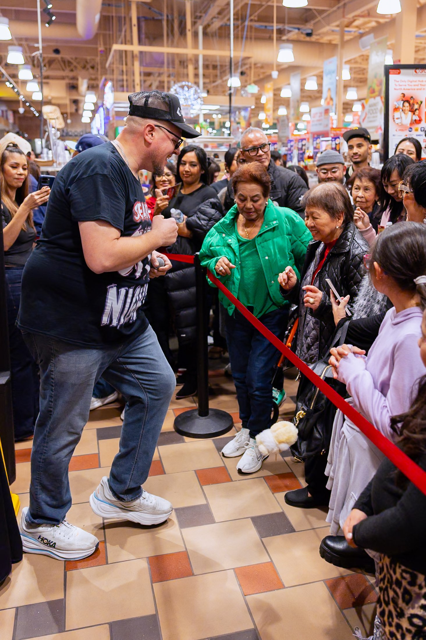 A man with a microphone engaging with a crowd of diverse women and girls in Seafood City, some smiling and taking photos, with bright overhead lights and store displays in the background.