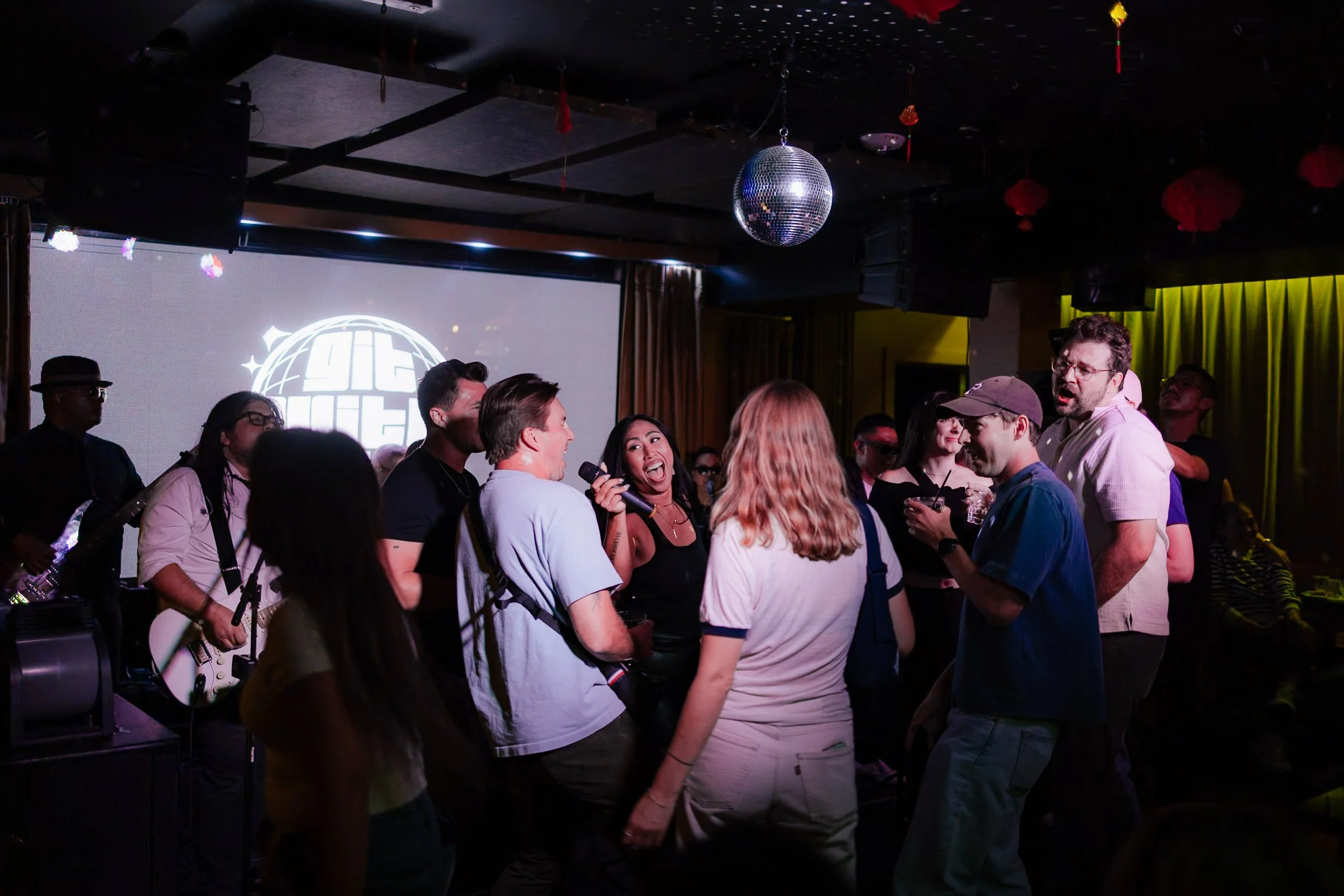 People dancing and singing at a nightclub with a band playing in the background and a disco ball hanging from the ceiling.