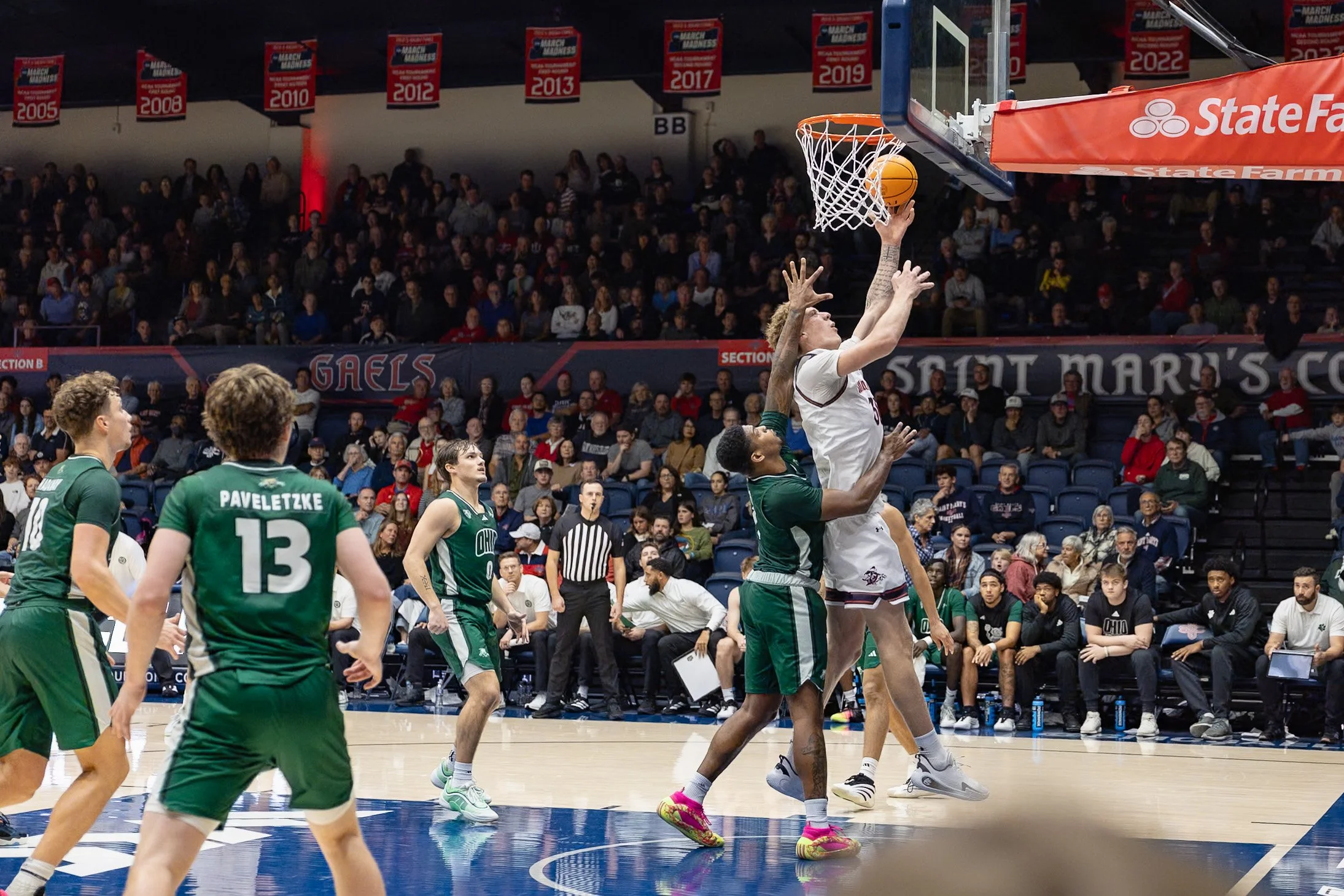 A basketball game with players near the hoop; a player in a white uniform jumps to shoot while a player in a green uniform blocks. The crowd watches from the stands.
