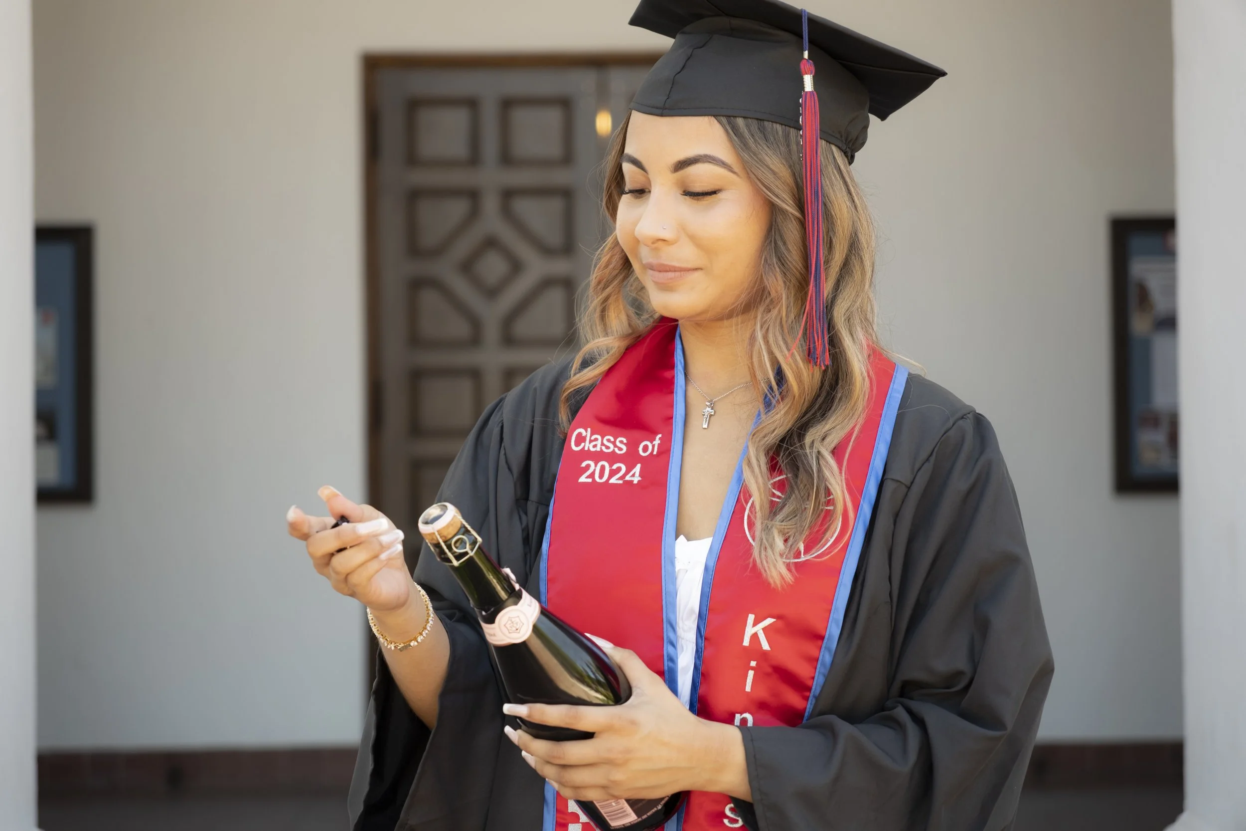 Young woman in graduation cap and gown holding a champagne bottle and opening it at graduation ceremony.