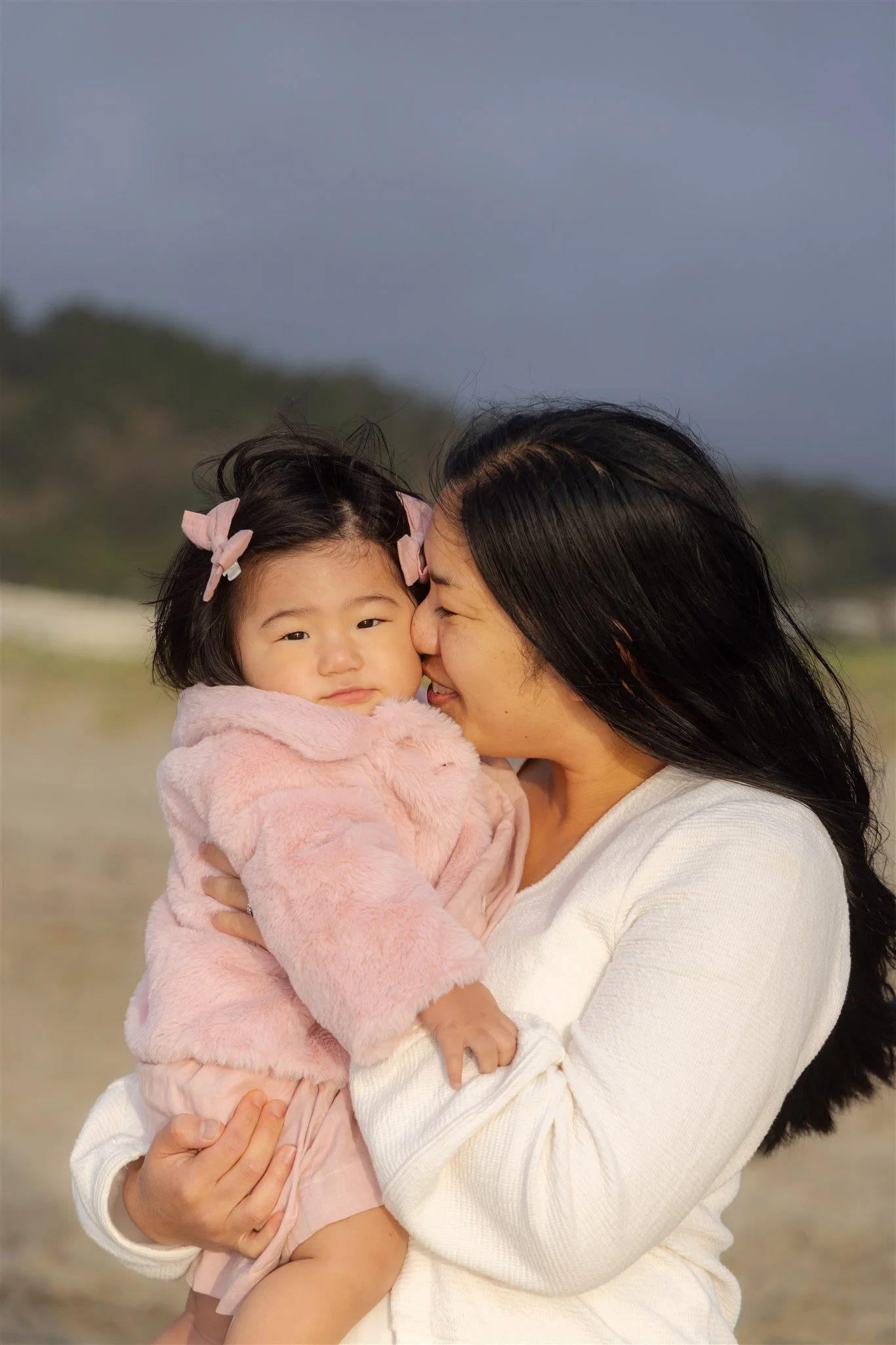 A woman is holding a young girl close as the woman kisses the girl on the cheek outdoors, with hills and cloudy sky in the background.