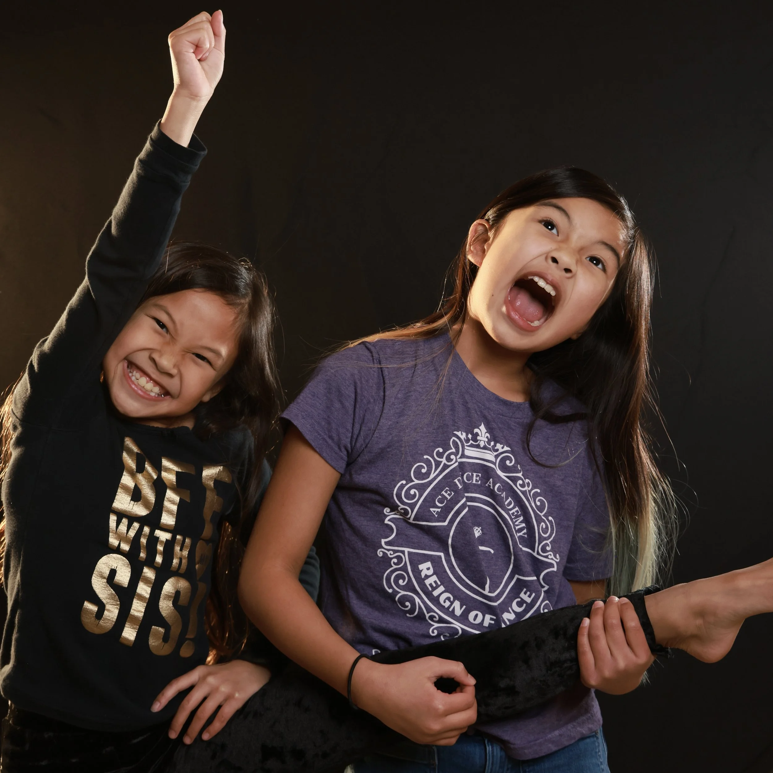 Two young girls smiling and laughing, one raising her hand with a fist, with dark background.