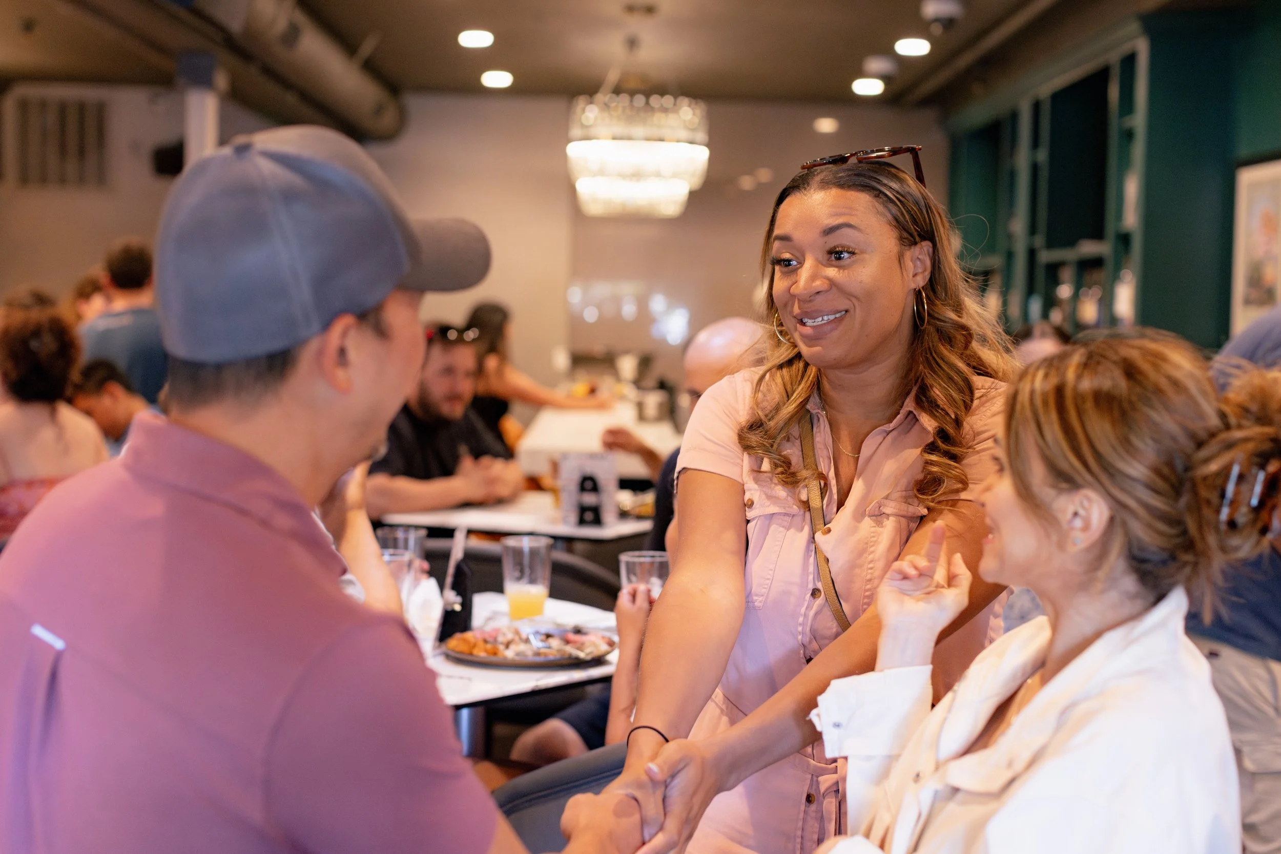 A woman is smiling and shaking hands with a man at a social event, with other people in the background.