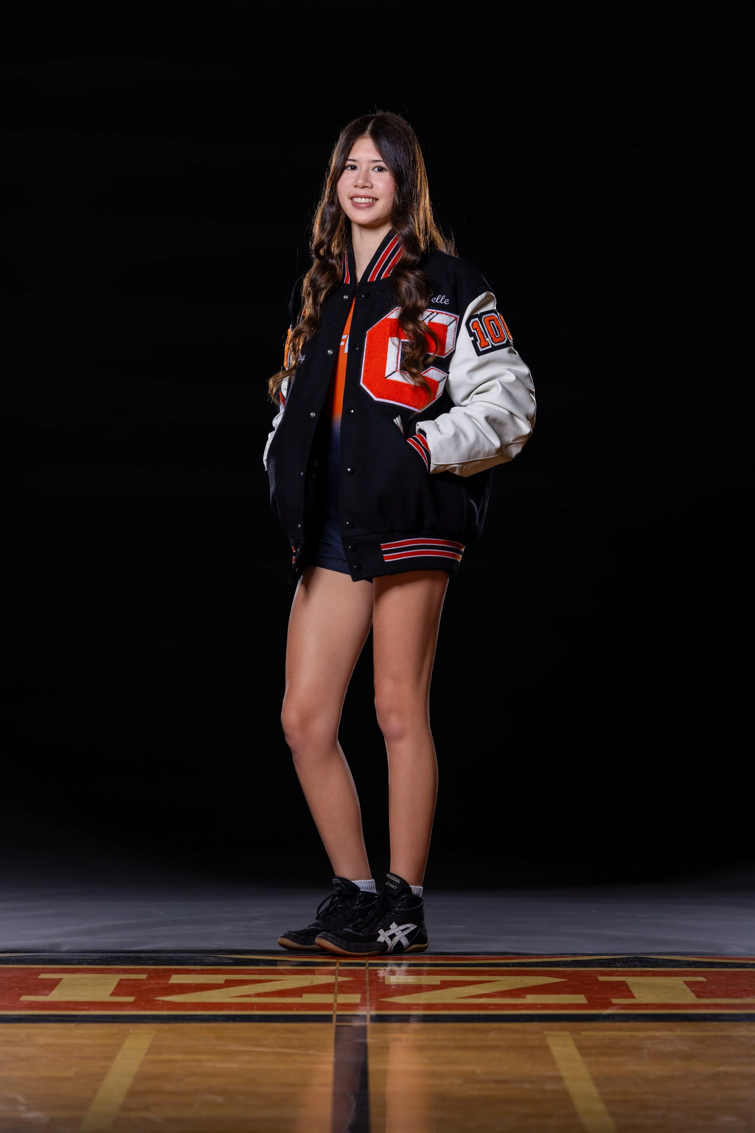 A young woman standing on a basketball court wearing a varsity jacket and athletic shoes, smiling at the camera.