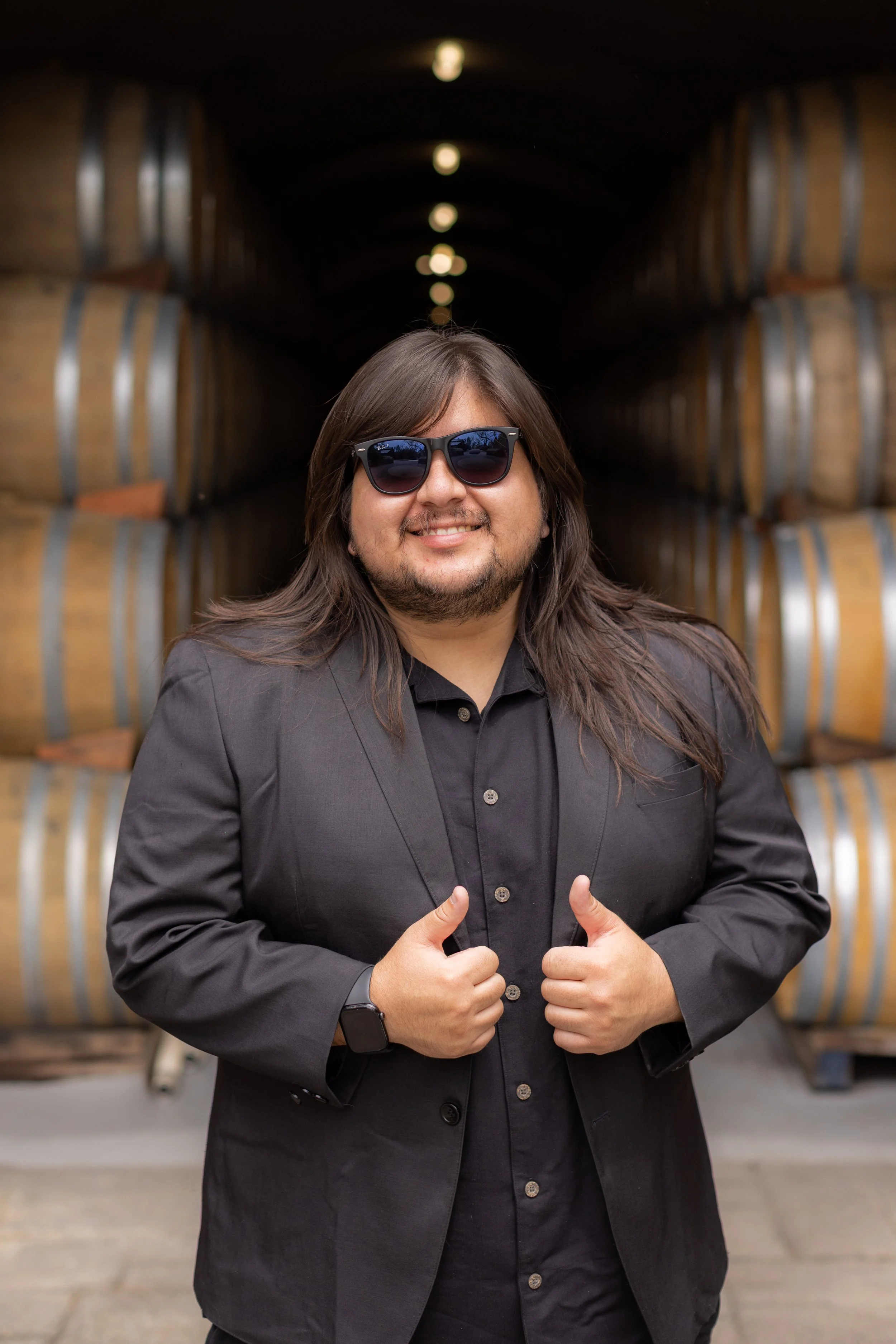 A man with long hair, sunglasses, and a black suit gives a thumbs-up in front of stacked wine barrels.