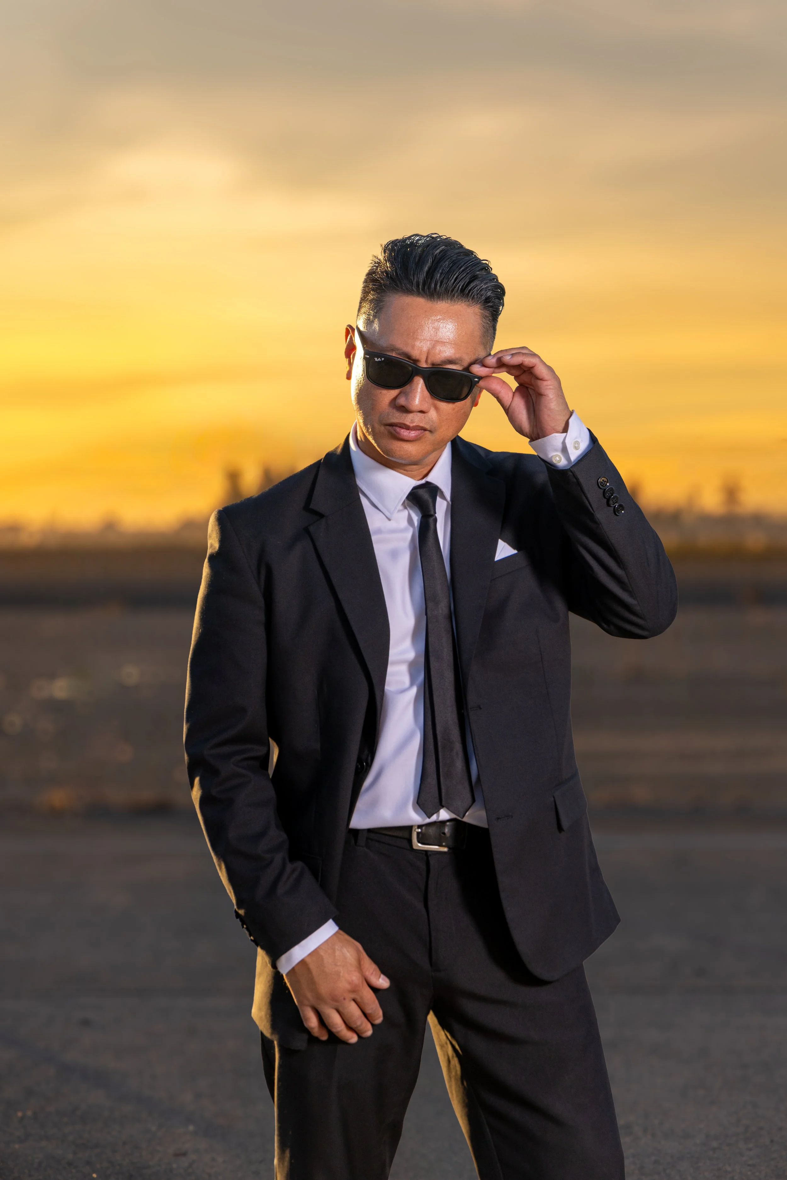 A man dressed in a black suit, white shirt, and black tie adjusting his sunglasses at sunset.