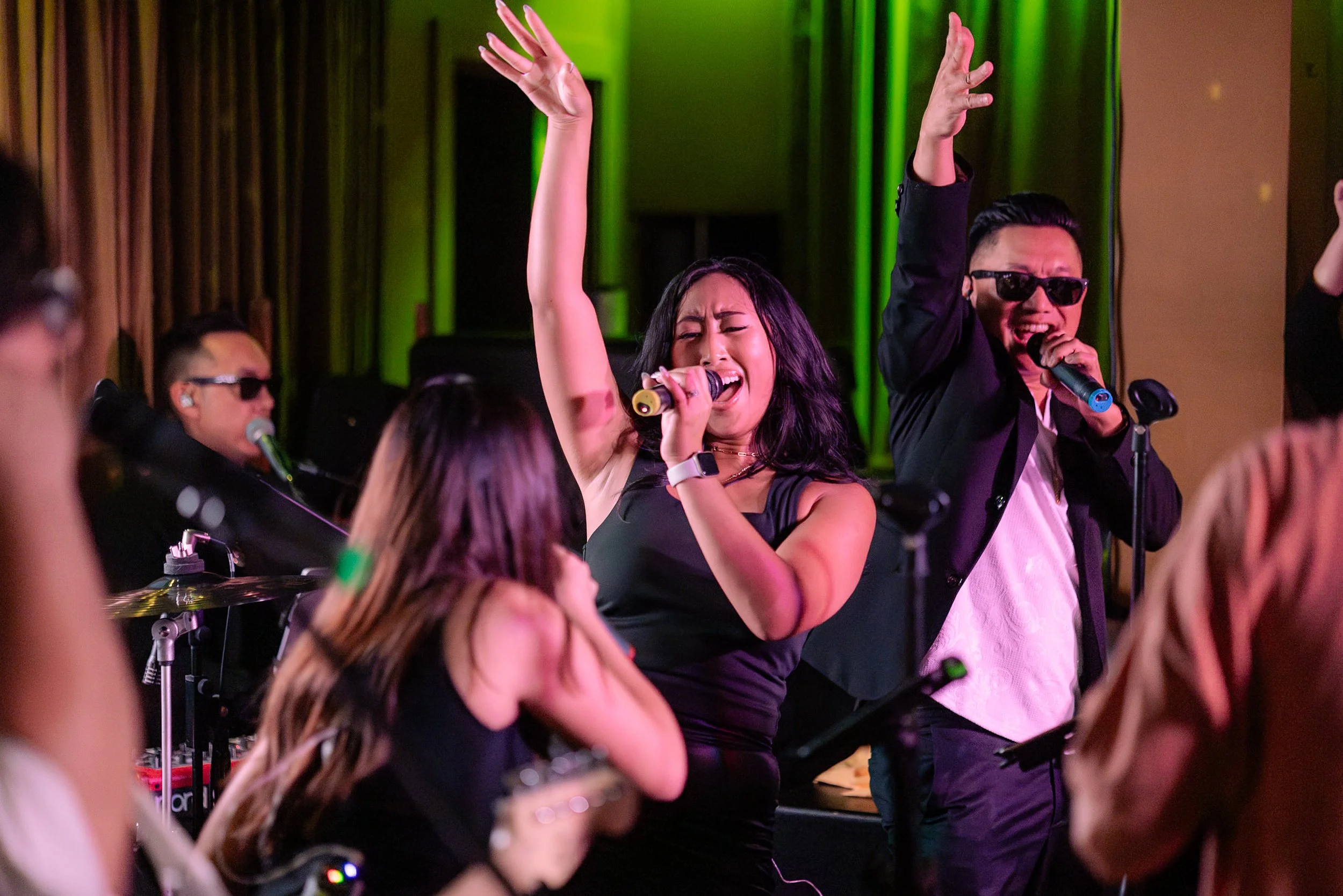 A group of people singing and dancing at a lively event, with a woman in the center holding a microphone and raising her hand, and a man beside her singing into a microphone, both wearing sunglasses. The scene is vibrant with colorful lighting and a 