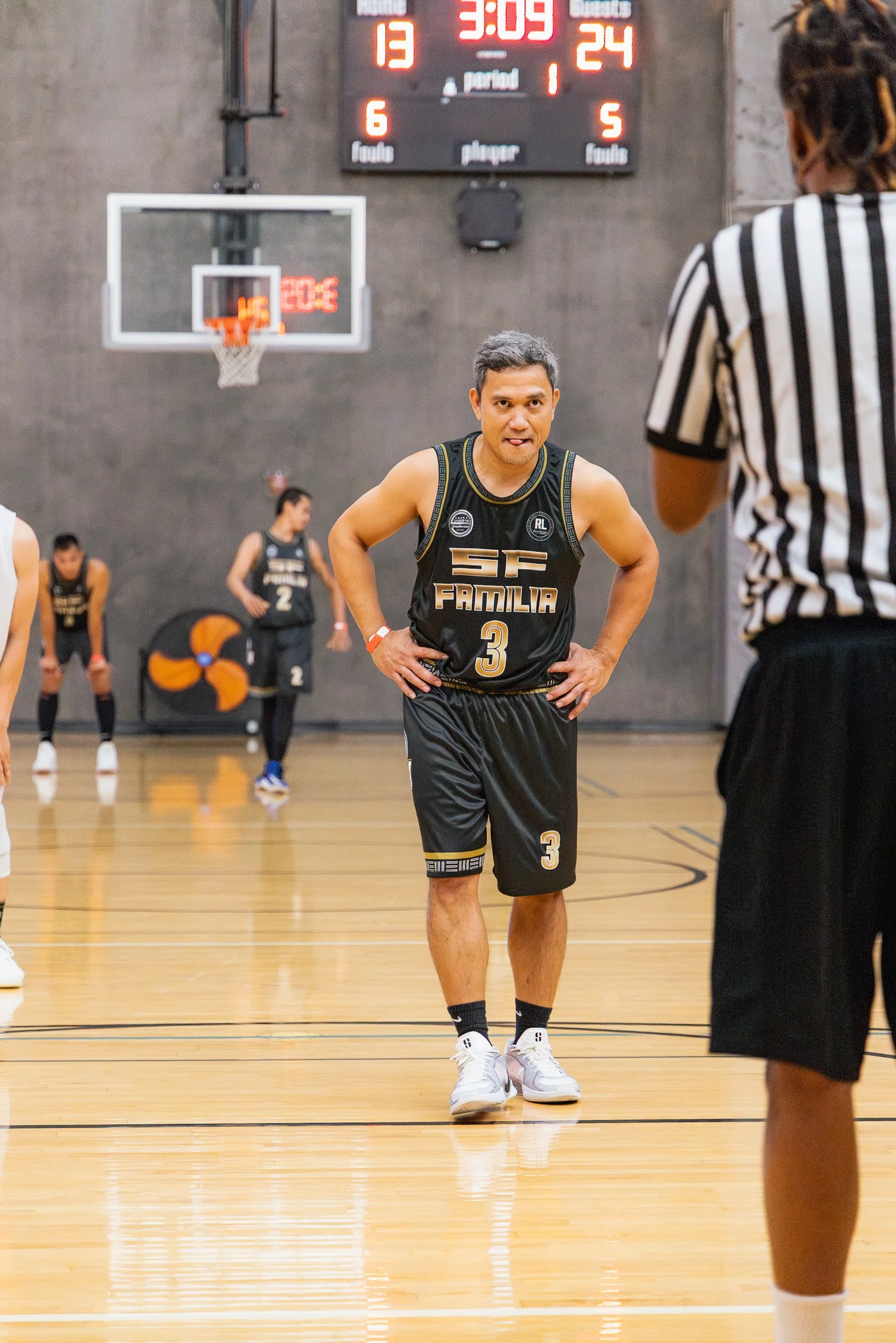 A man in a black basketball uniform with the words SF Familia and the number 3 is standing on a basketball court, looking at a referee. There are other players in the background and a scoreboard above showing the score as 13-24 with 3:09 remaining in