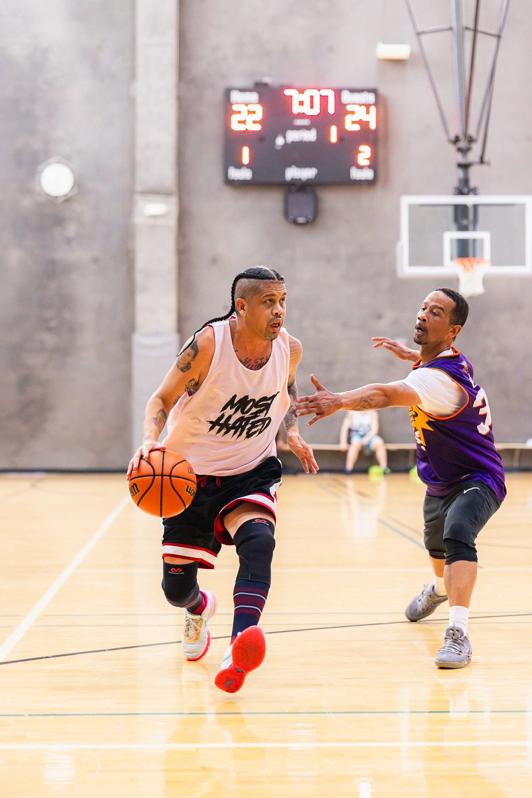 Two men playing basketball in an indoor gym, one dribbling the ball while the other defends. A basketball hoop is visible in the background. The scoreboard above shows the score as 22-24 with 7:07 remaining in the game.