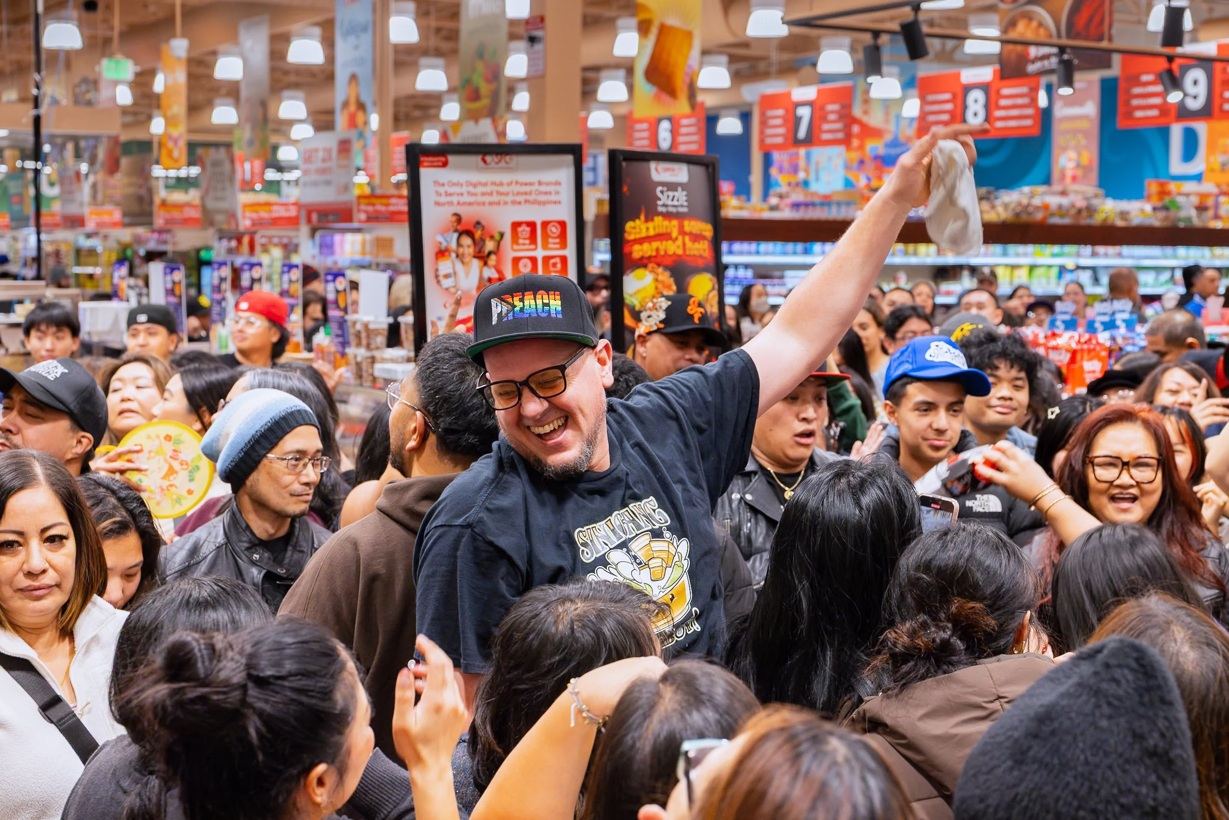 A man smiling and raising his hand in a crowded store or event, surrounded by many people.