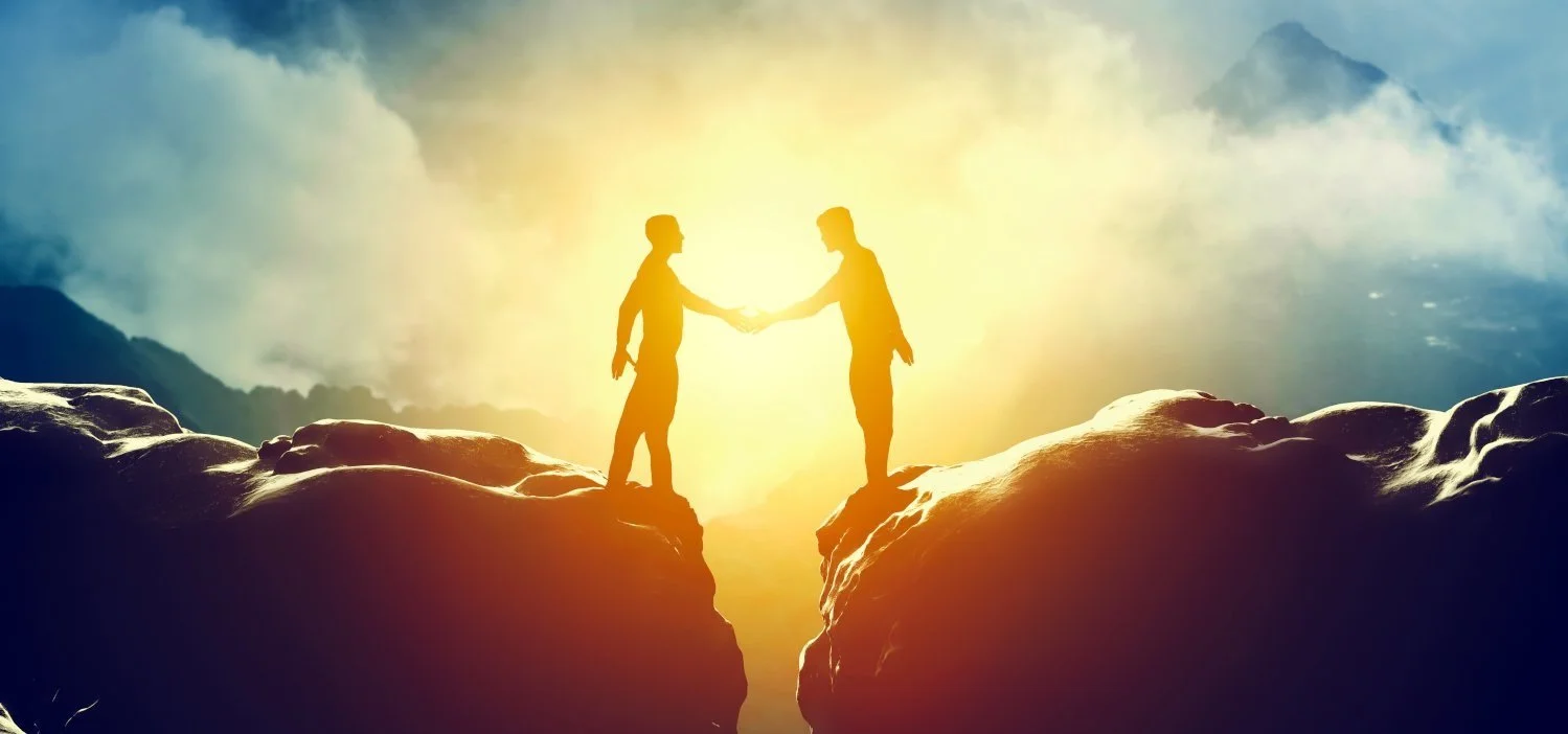 Two silhouetted people shaking hands on top of separate rocky cliffs at sunset or sunrise, with mountains and clouds in the background.