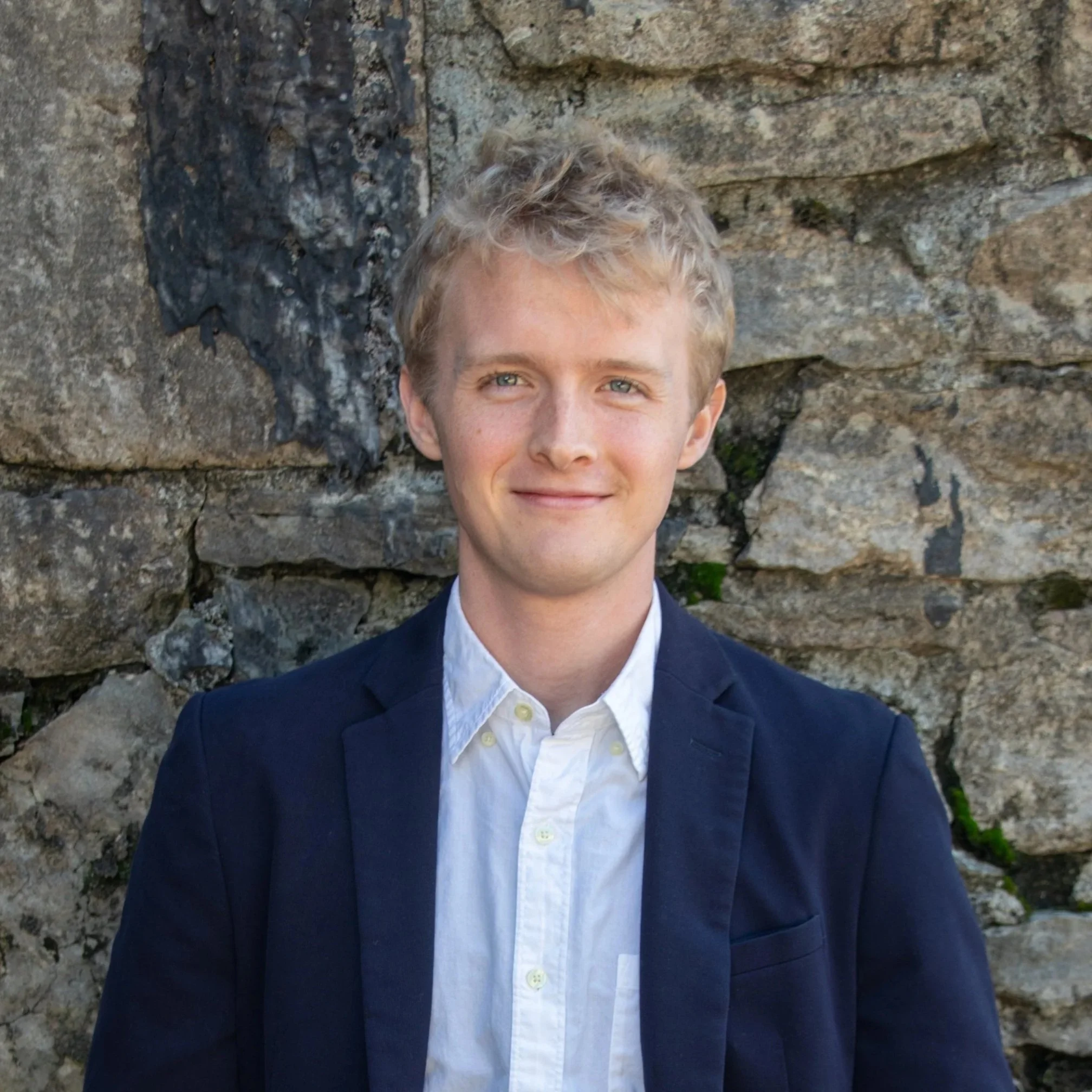 A young man with light curly hair and green eyes wearing a navy blue blazer and white shirt stands in front of a stone wall.