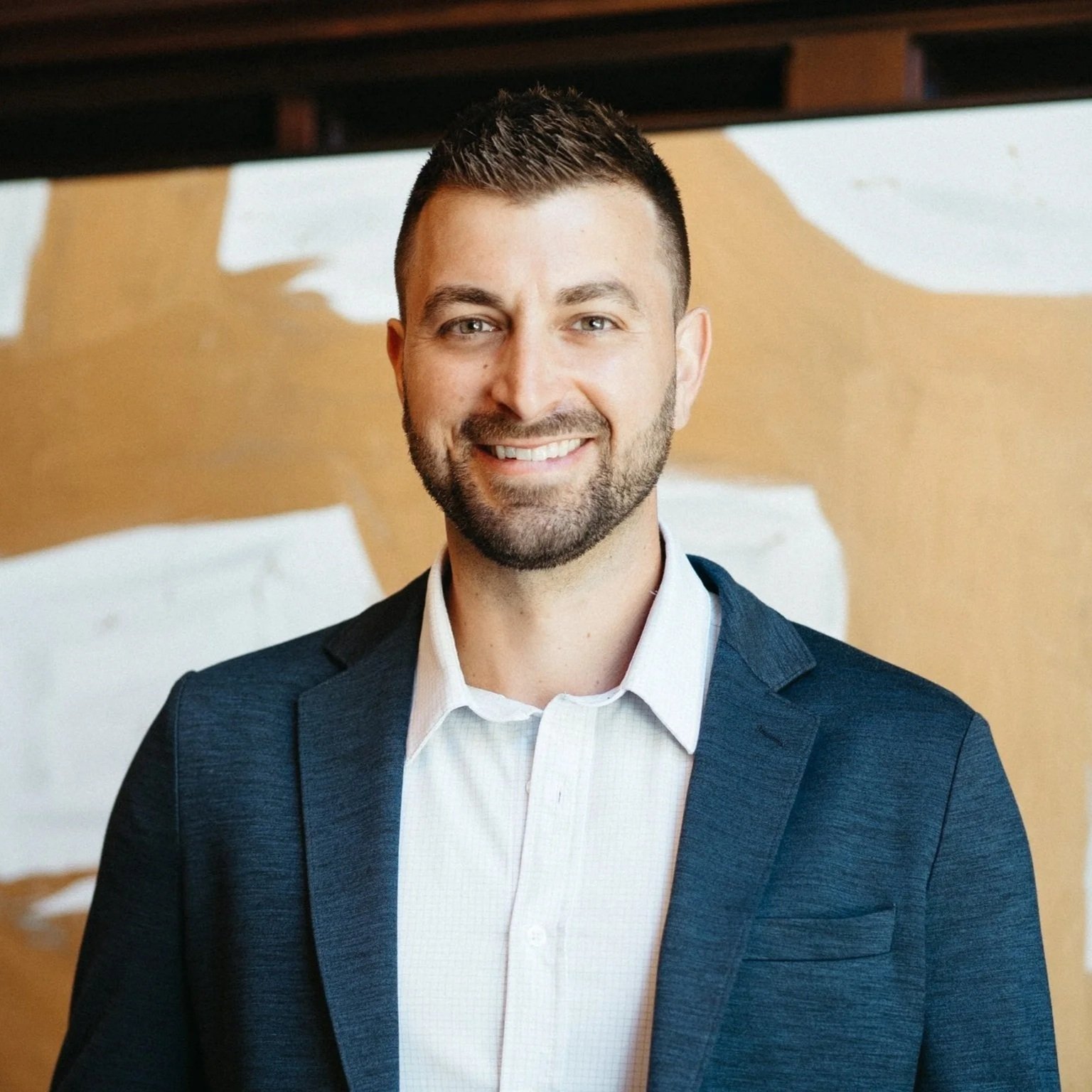 Portrait of a smiling man with short dark hair, beard, blue suit, and white shirt, in front of a wooden and white background.