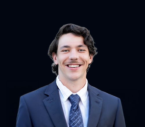 A young man wearing a navy blue suit, white shirt, and patterned tie, smiling against a dark background.