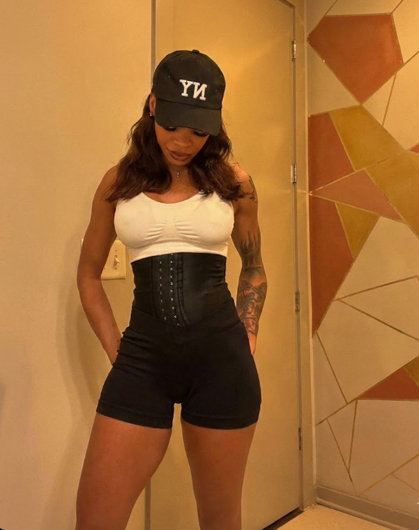 A woman wearing a black hat with 'NY' on it, a white top, a black waist trainer, and black shorts standing in front of a geometric patterned wall.